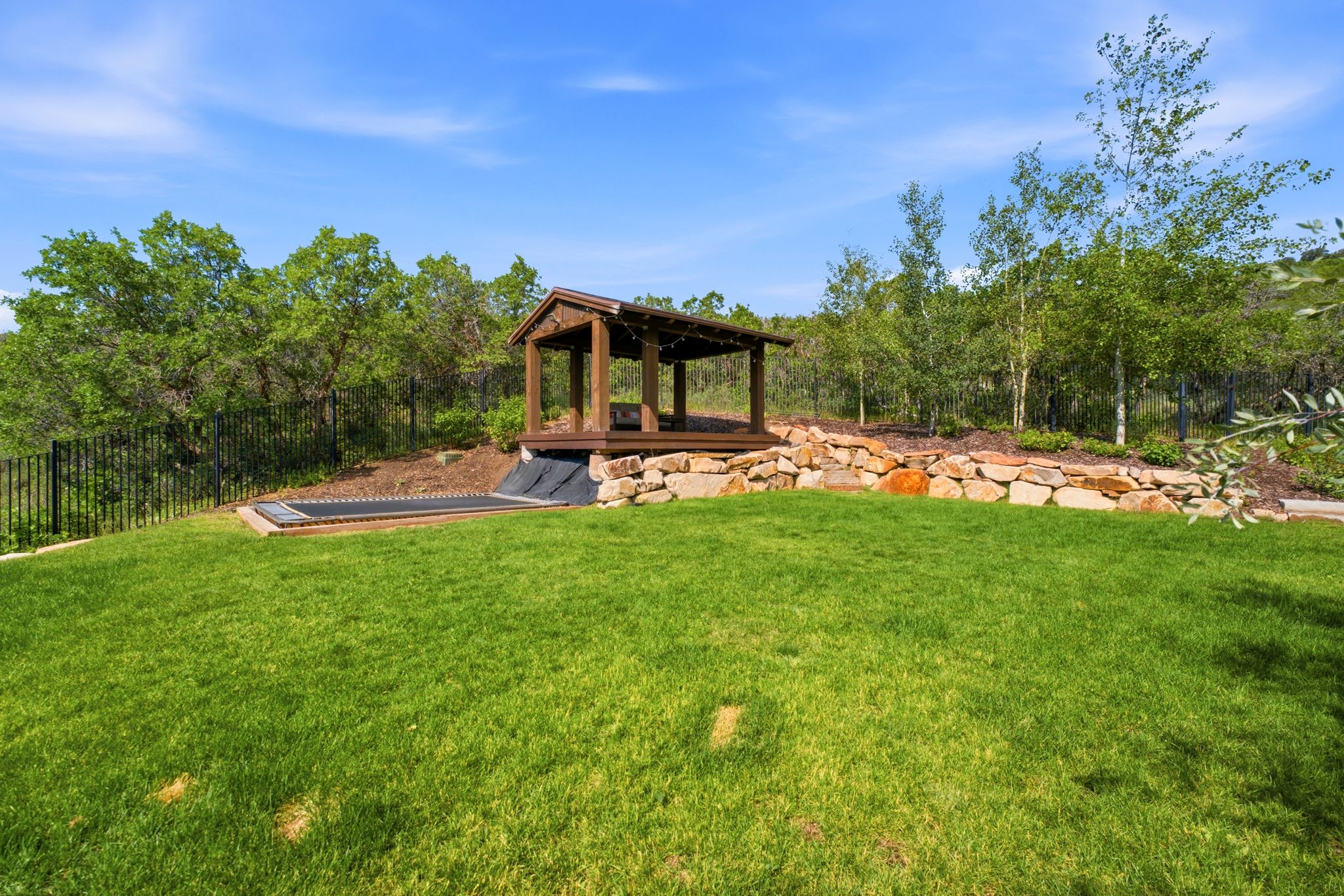 Expansive lawn area featuring a gazebo with a wood-finish structure and integrated lighting, a built-in trampoline, a natural stone retaining wall, and mature trees