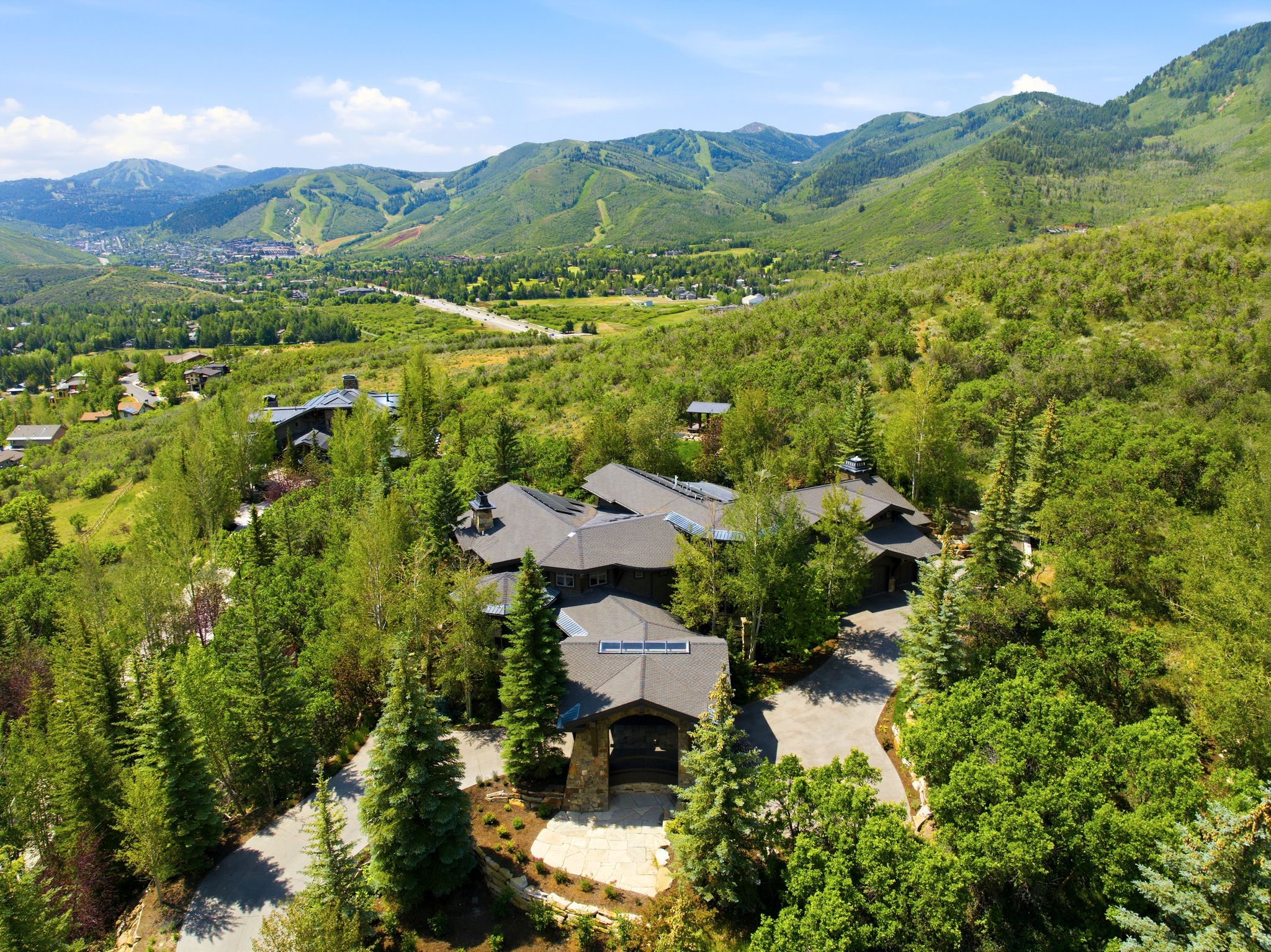 Expansive mountain estate featuring multiple dark-toned structures with sloped roofs, surrounded by dense green trees and foliage