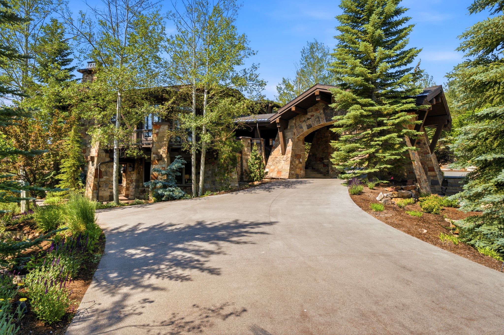 Stone-clad residence featuring an arched porte-cochère, a winding paved driveway, and mature evergreen trees