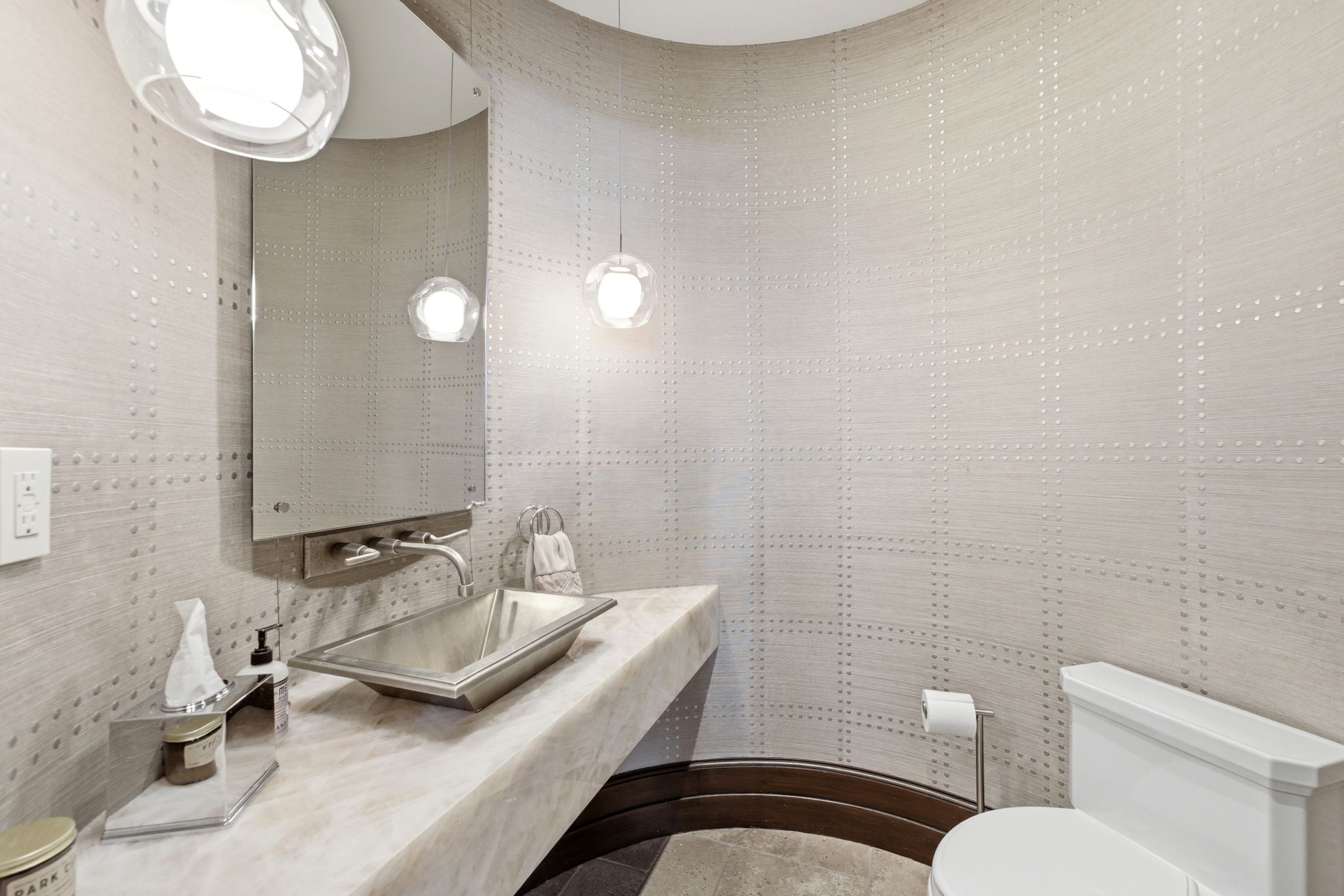 Powder room featuring a curved wall with textured wallcovering, a rectangular vessel sink set on a stone countertop, a wall-mounted faucet, and two pendant light fixtures