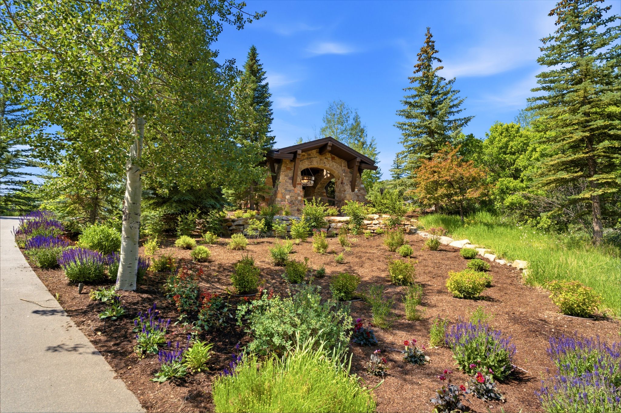 Stone-clad entrance structure featuring a timber roof, surrounded by mature trees, varied shrubs, and perennial plantings