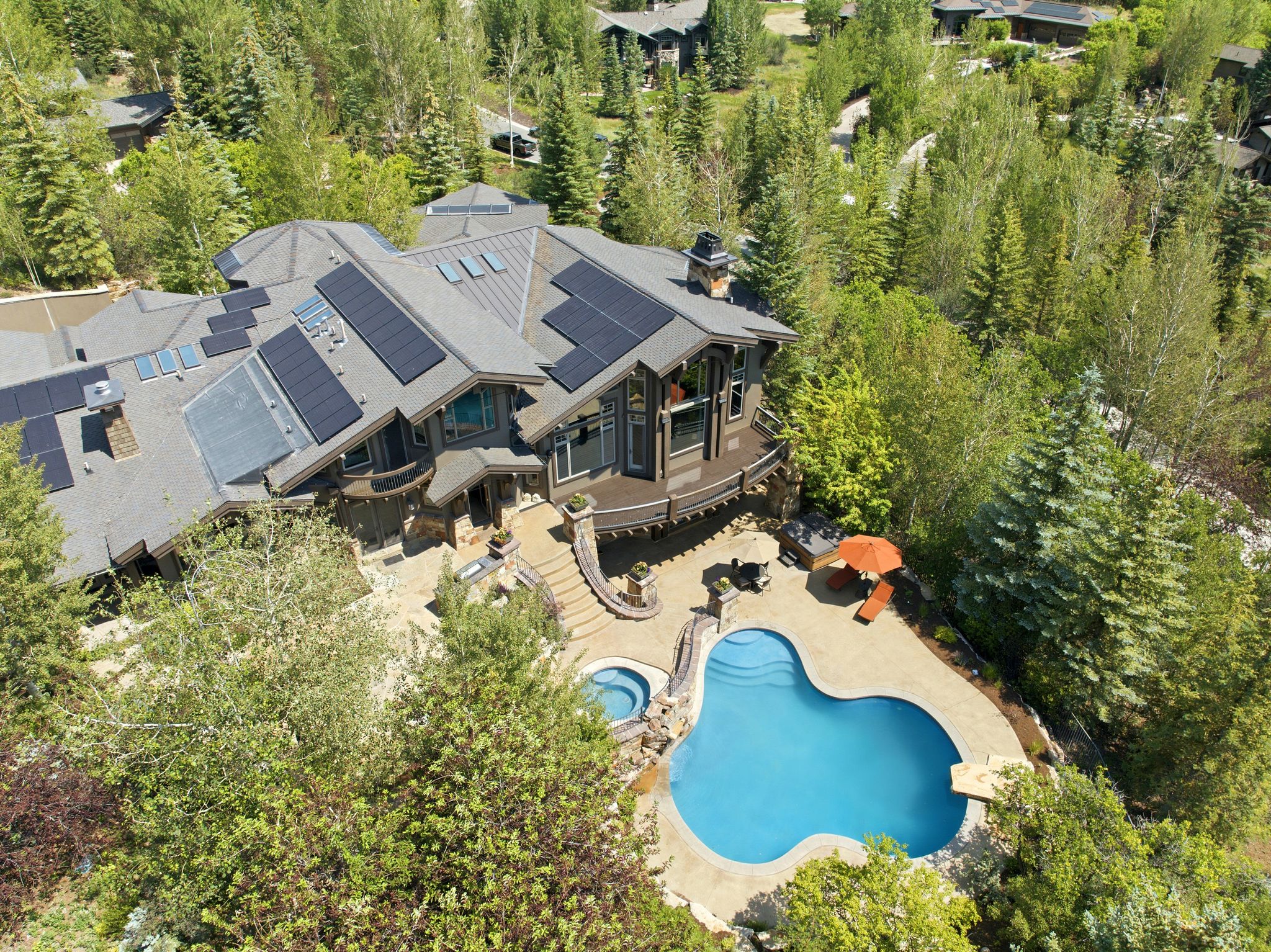 Contemporary residence featuring a multi-level roofline with integrated solar panels, an expansive outdoor patio, a freeform swimming pool, and an in-ground spa, all surrounded by dense mature trees