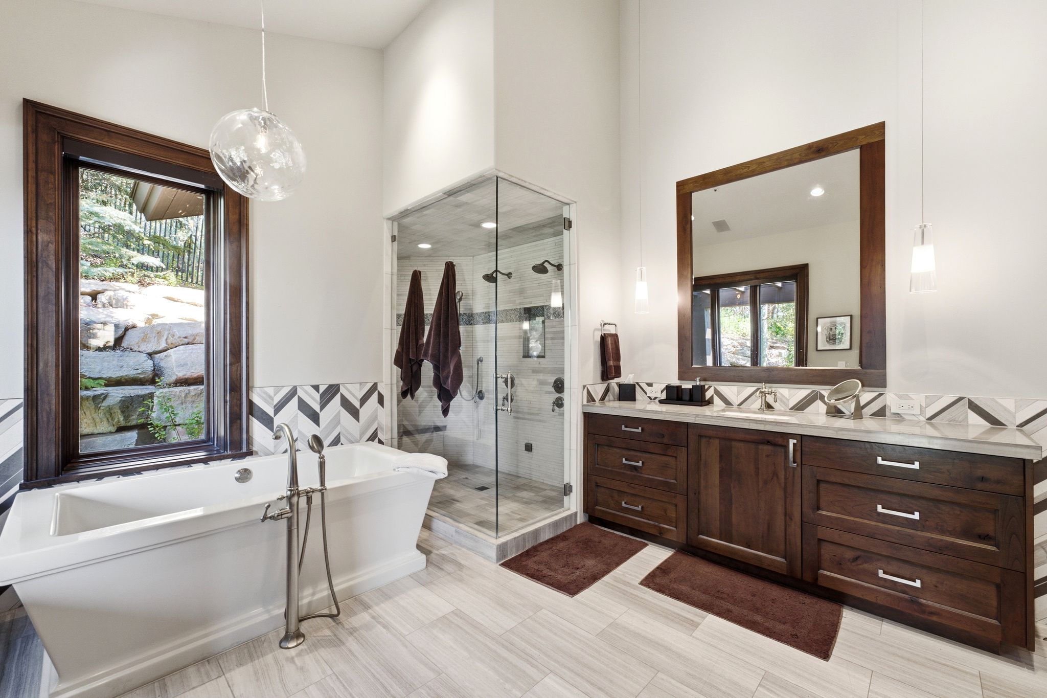 Bathroom featuring a freestanding soaking tub with floor-mounted faucet, a glass-enclosed shower, a double vanity with stone countertop, and herringbone-patterned tile accents