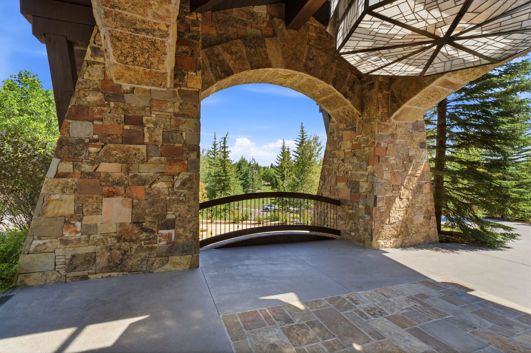 Stone archway entry featuring a patterned overhead light fixture, stone veneer columns, a concrete pathway, and a decorative metal railing bridge