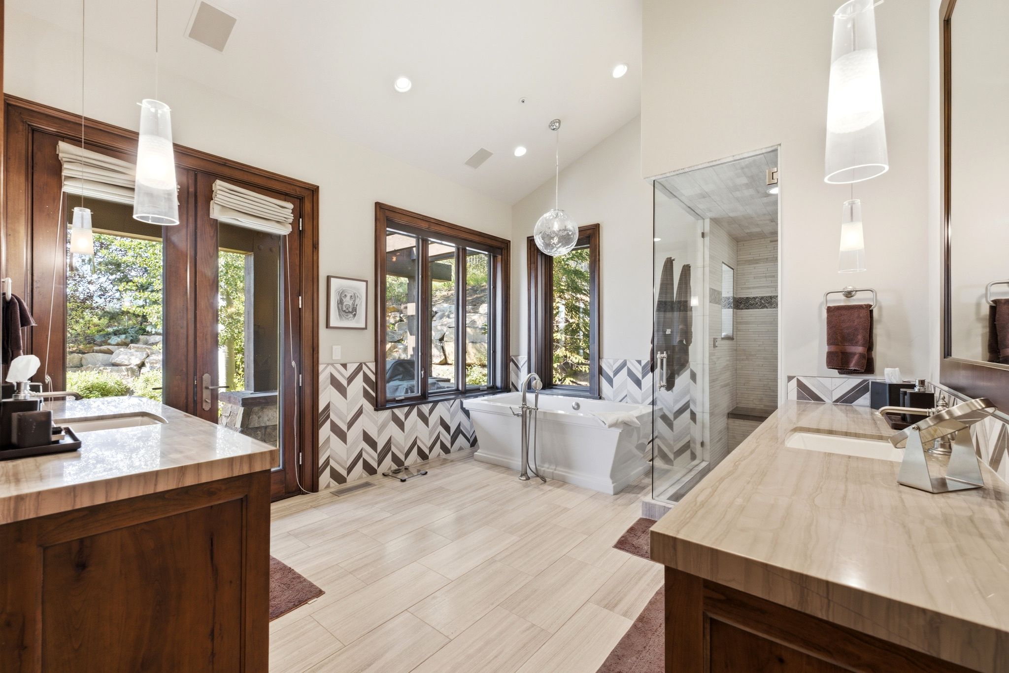Spacious bathroom featuring a freestanding tub, glass-enclosed shower, two wood vanities with integrated sinks, light-toned flooring, and chevron-patterned wall tiling