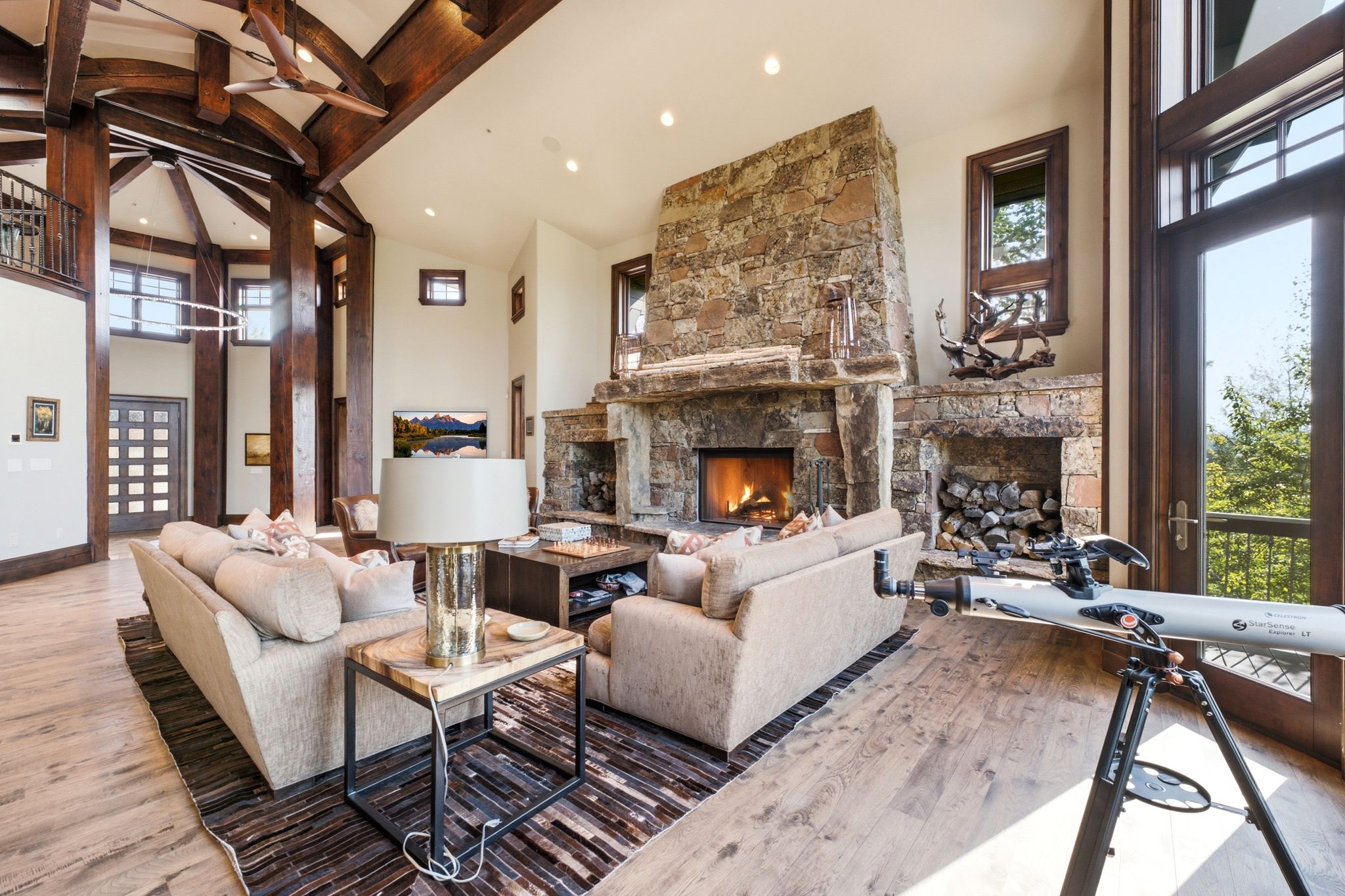 Spacious living area featuring a prominent stone fireplace, vaulted ceilings with exposed wood beams, wood-finish flooring, and a full-height glass door
