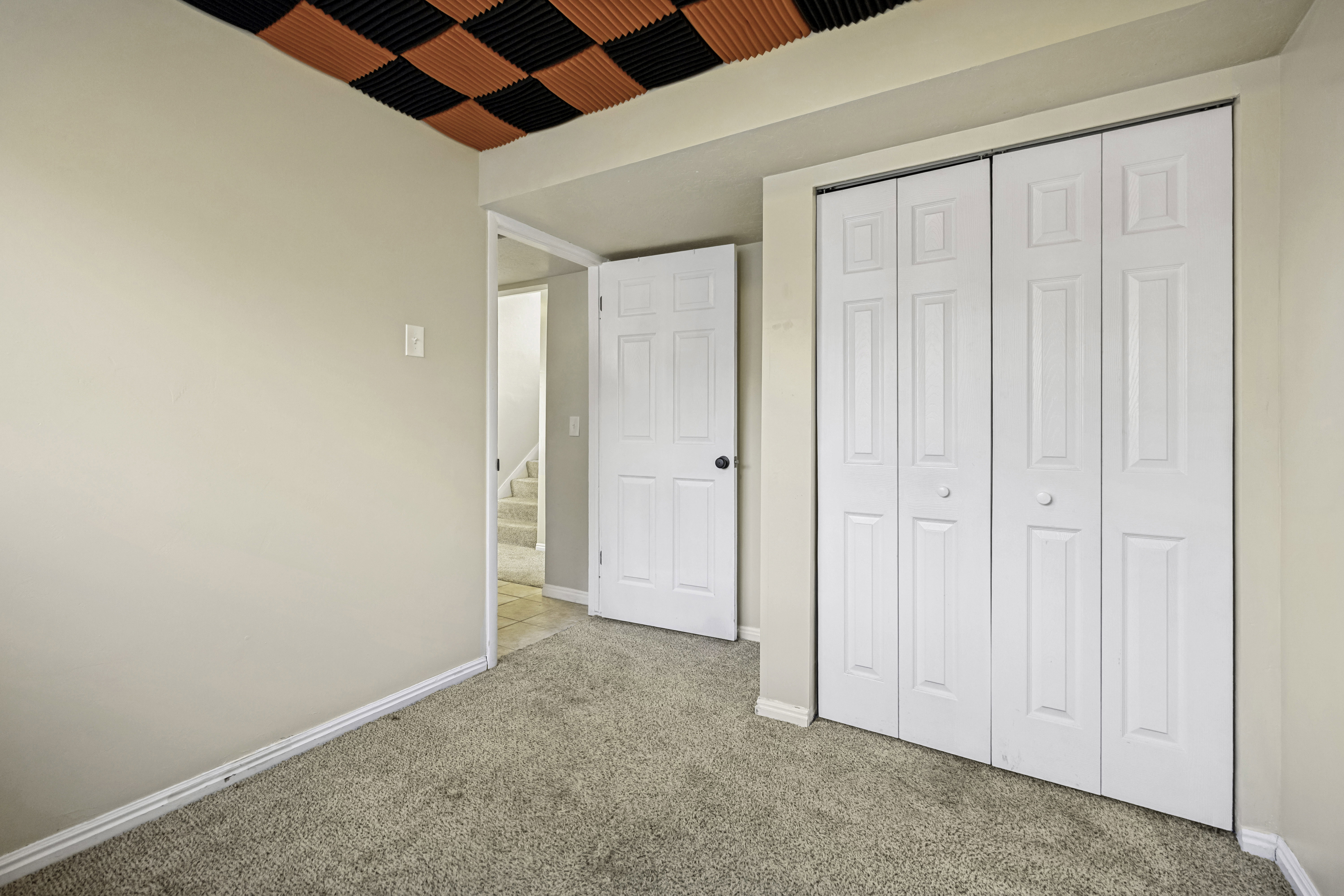 Room featuring neutral carpet flooring, light beige wall paint, a bi-fold closet with panel doors, and a ceiling with black and orange acoustic panels