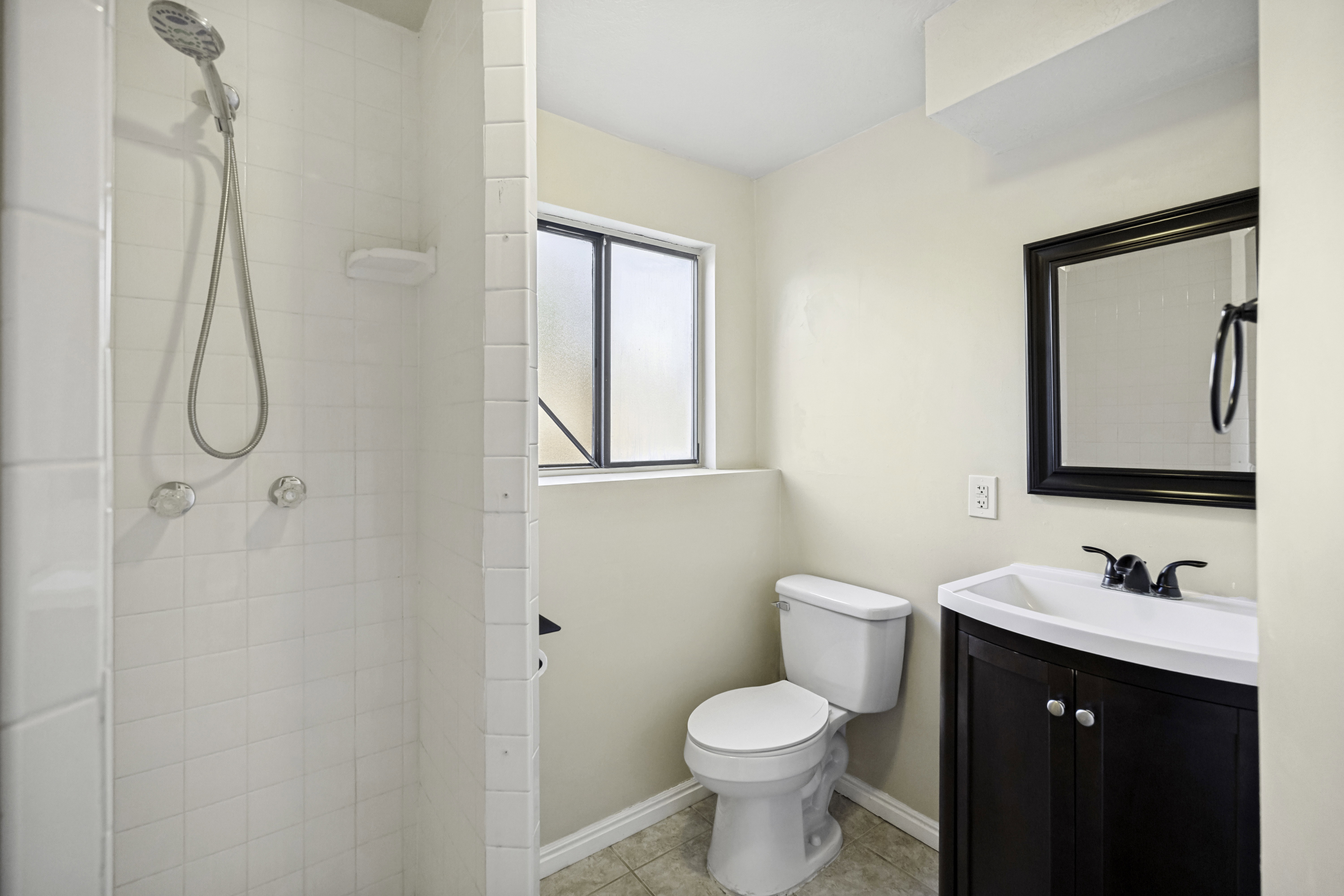 Bathroom featuring a walk-in shower with white tile surround, a dark wood-finish vanity with an integrated sink, a dark framed mirror, and a window with obscured glass