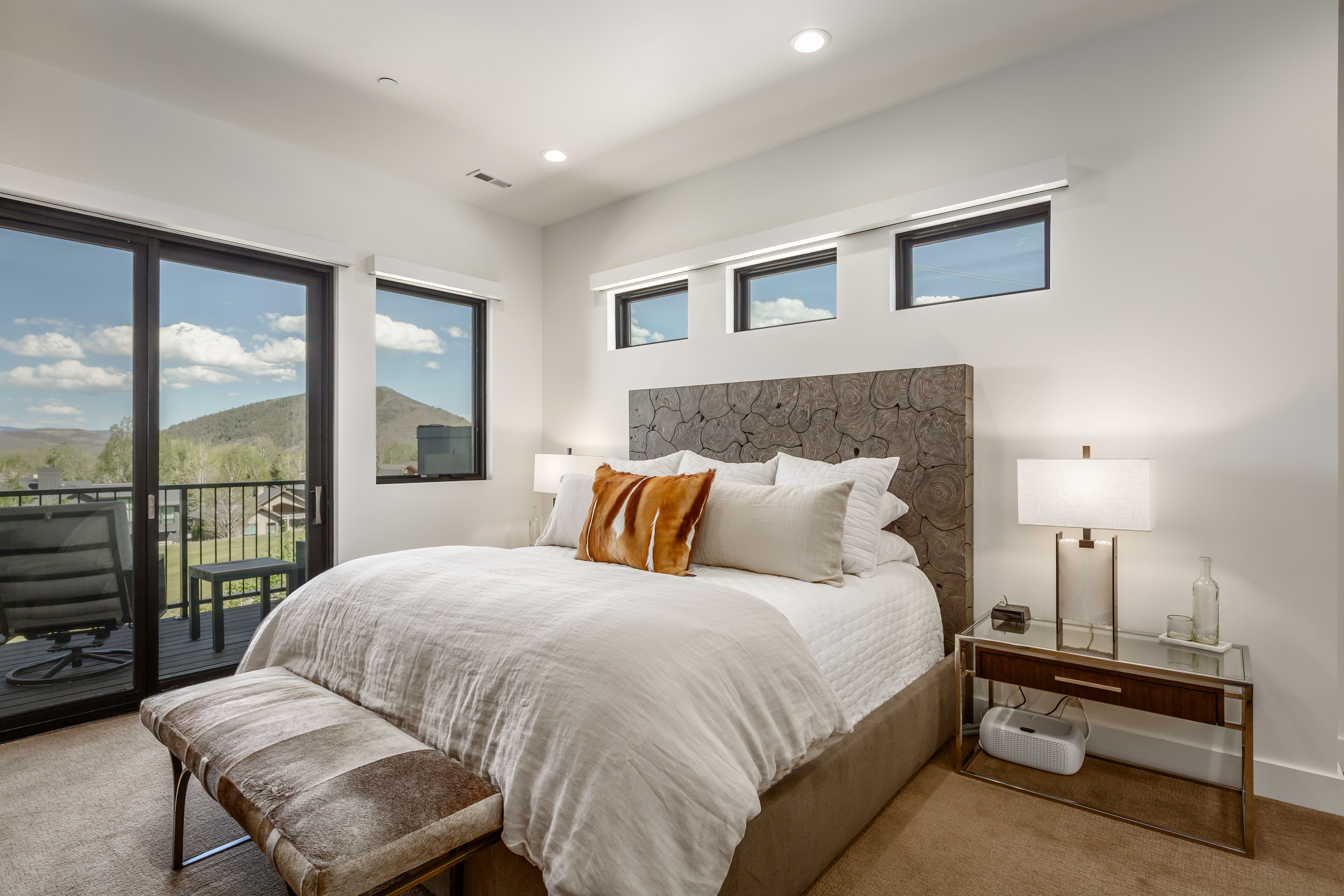 Spacious room featuring a private balcony with black railings, a large sliding glass door, three horizontal transom windows, recessed lighting, and a distinctive wood-textured headboard