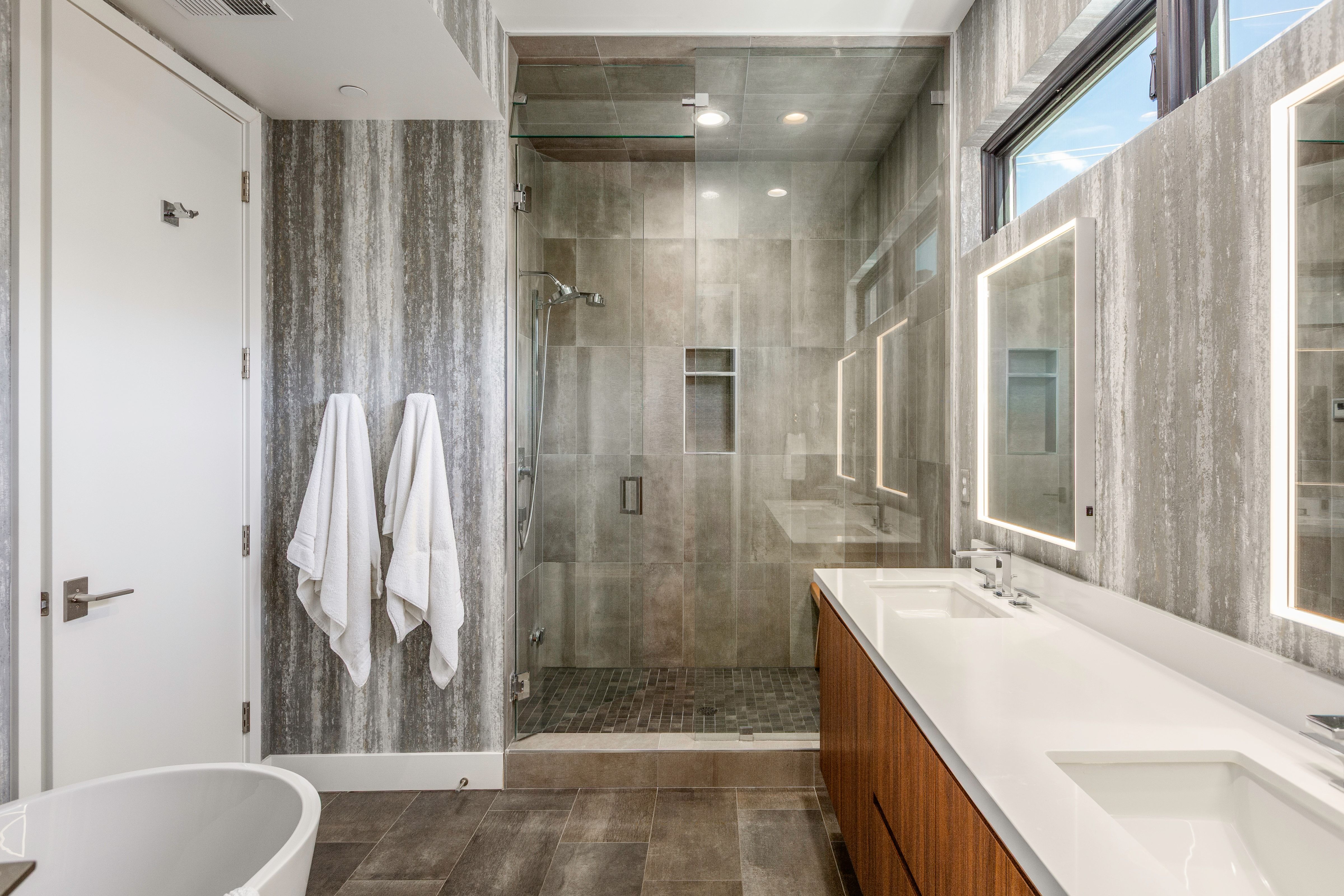 Contemporary bathroom featuring a spacious walk-in shower with a glass enclosure, dual vanity with undermount sinks and illuminated mirrors, a freestanding soaking tub, and wood-finish flooring