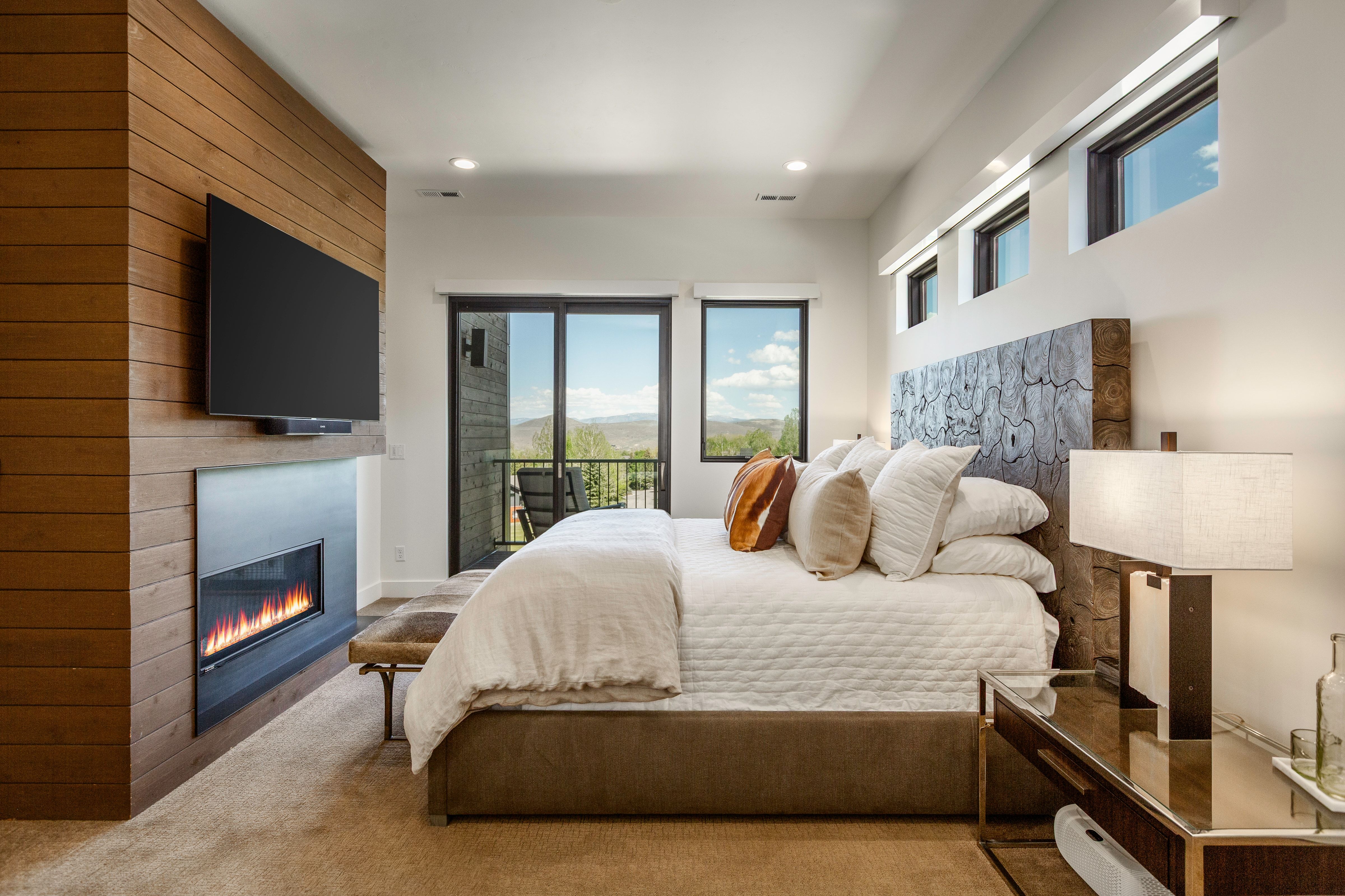 Modern bedroom featuring a wood-panel accent wall with an integrated linear fireplace, a flat-screen television, and expansive sliding glass doors opening to a balcony
