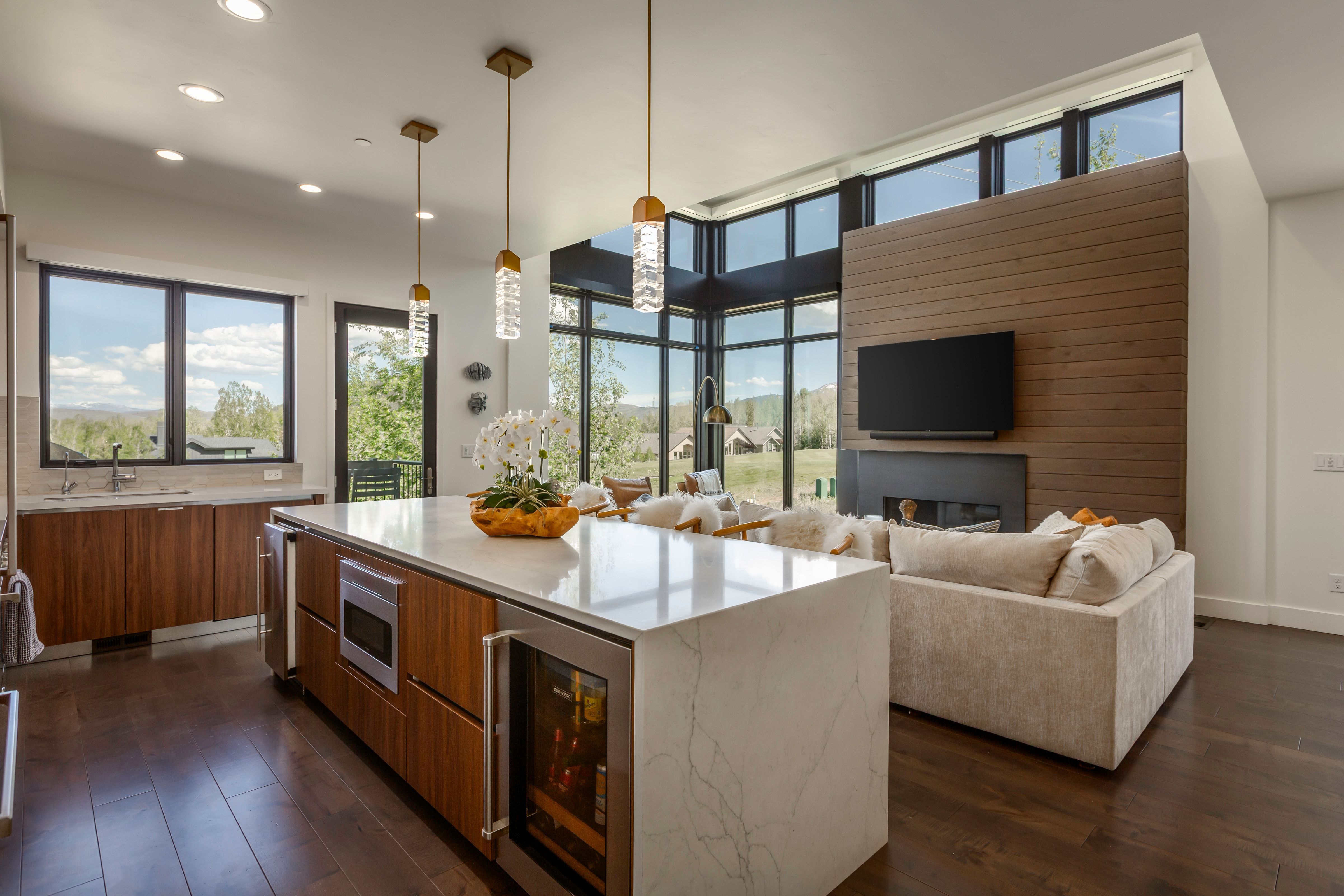 Spacious open-concept living area featuring an expansive kitchen island with a built-in beverage cooler and microwave, rich wood-finish flooring throughout, and a contemporary fireplace with a wood-paneled accent wall