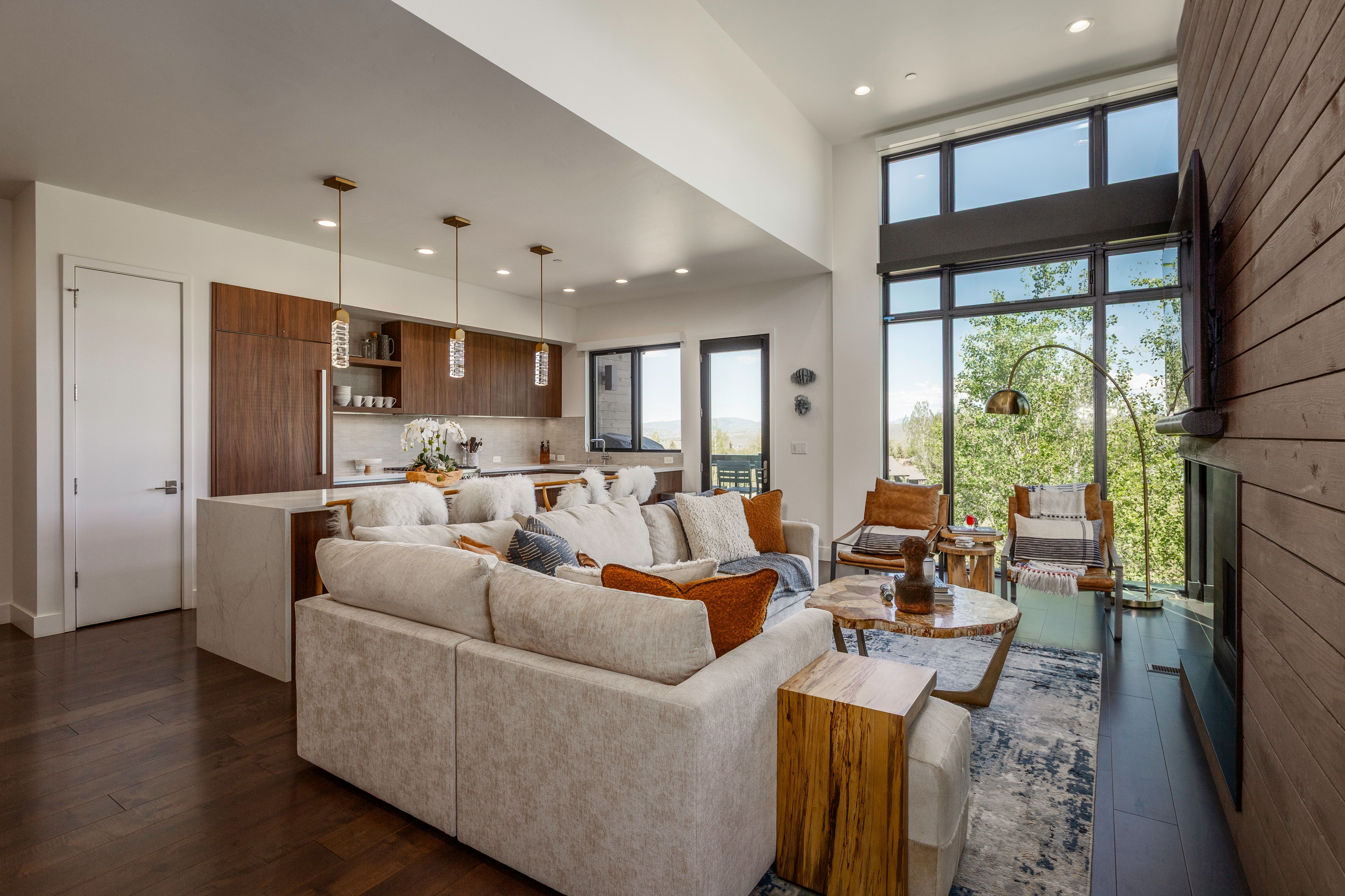 Open-concept living space featuring wood-finish flooring, a contemporary kitchen with dark wood cabinetry and integrated appliances, and a two-story window wall