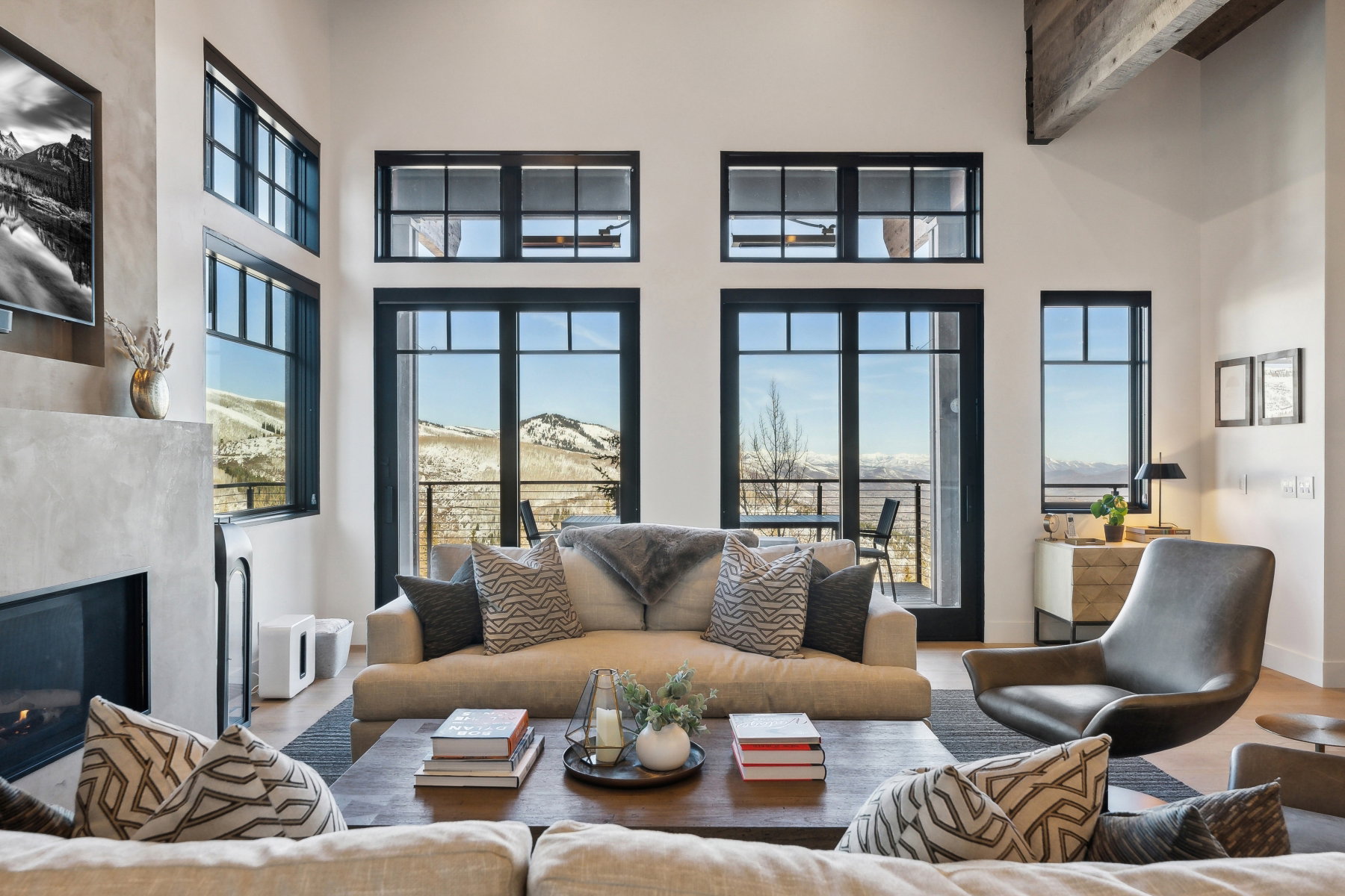 Double-height living space featuring dark-framed windows, a modern fireplace with a concrete-finish surround, exposed wood ceiling beams, and light-tone wood-finish flooring