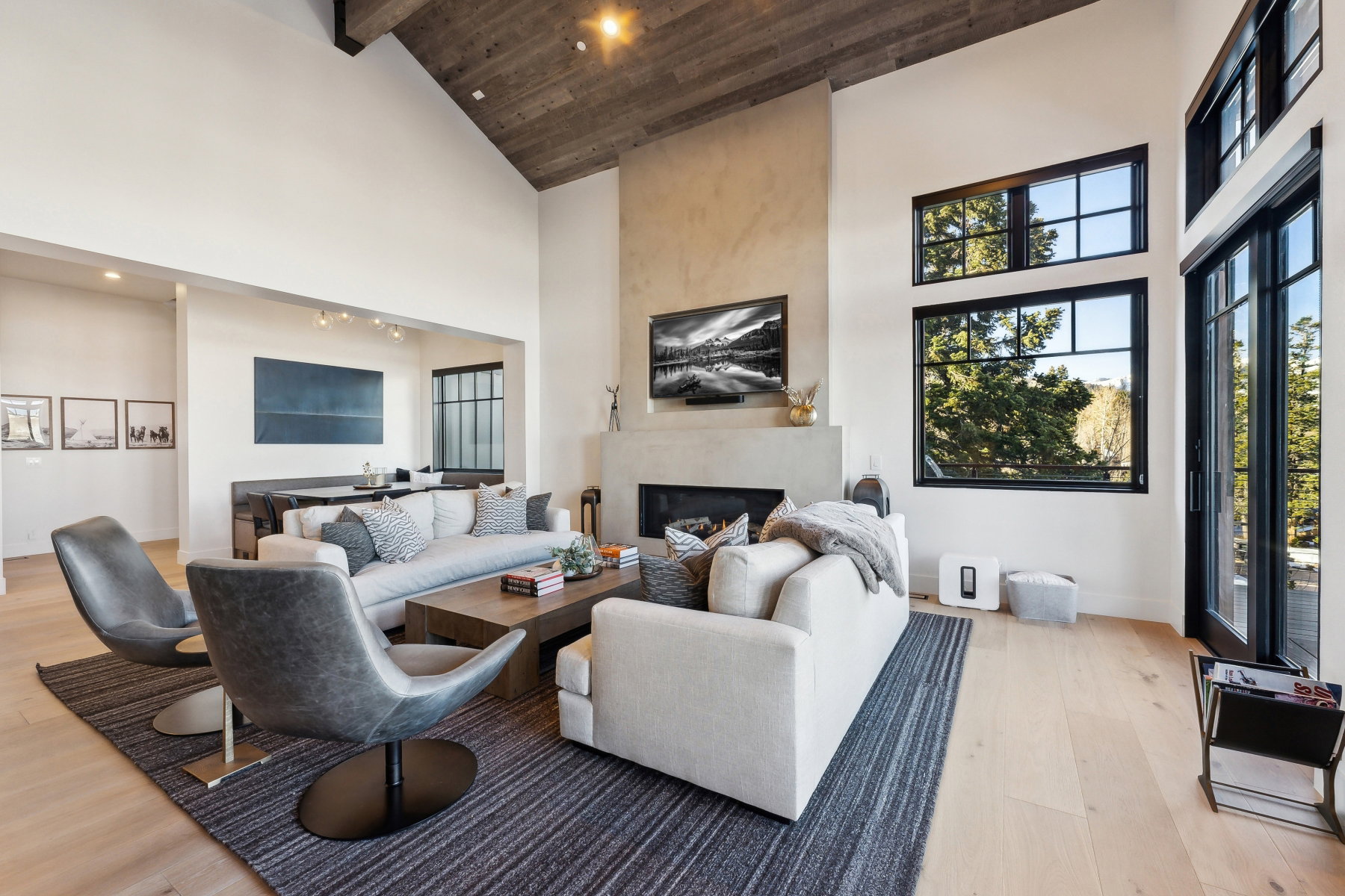 Vaulted ceiling with wood-finish planks, large format fireplace with concrete-finish surround, light wood-finish flooring, black-framed windows, and glass patio doors