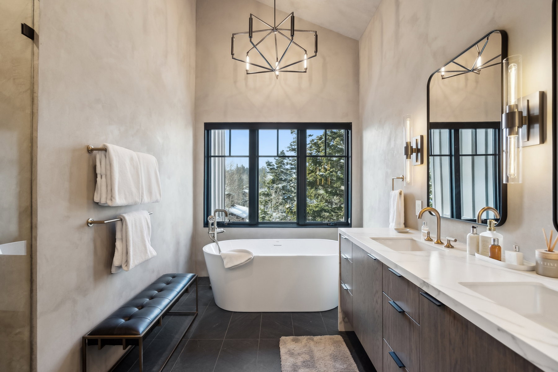 Bathroom featuring a freestanding soaking tub, a dual vanity with a light-toned countertop, a black-framed window, dark floor tiling, and a vaulted ceiling with a modern chandelier