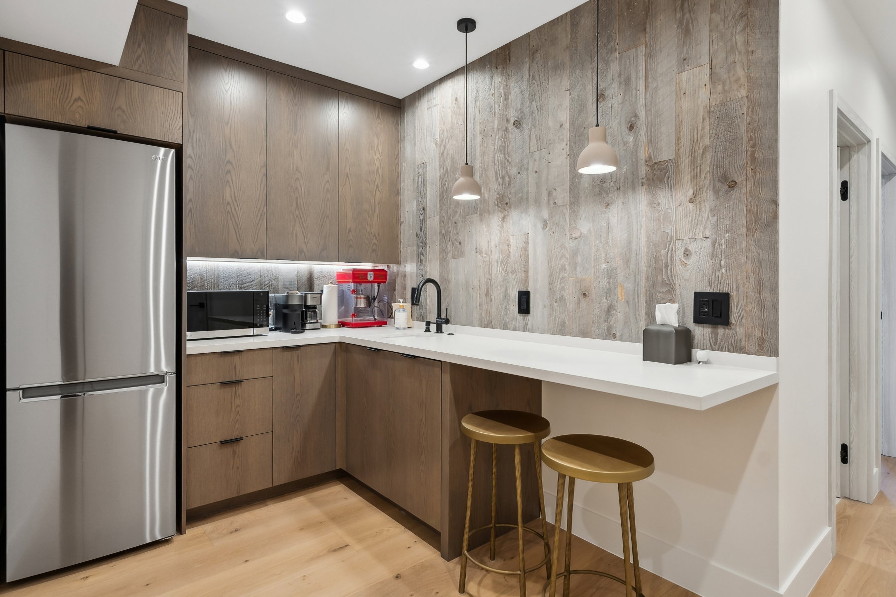 Compact kitchen space featuring custom wood cabinetry, stainless steel refrigerator, white countertops, and a distressed wood panel accent wall
