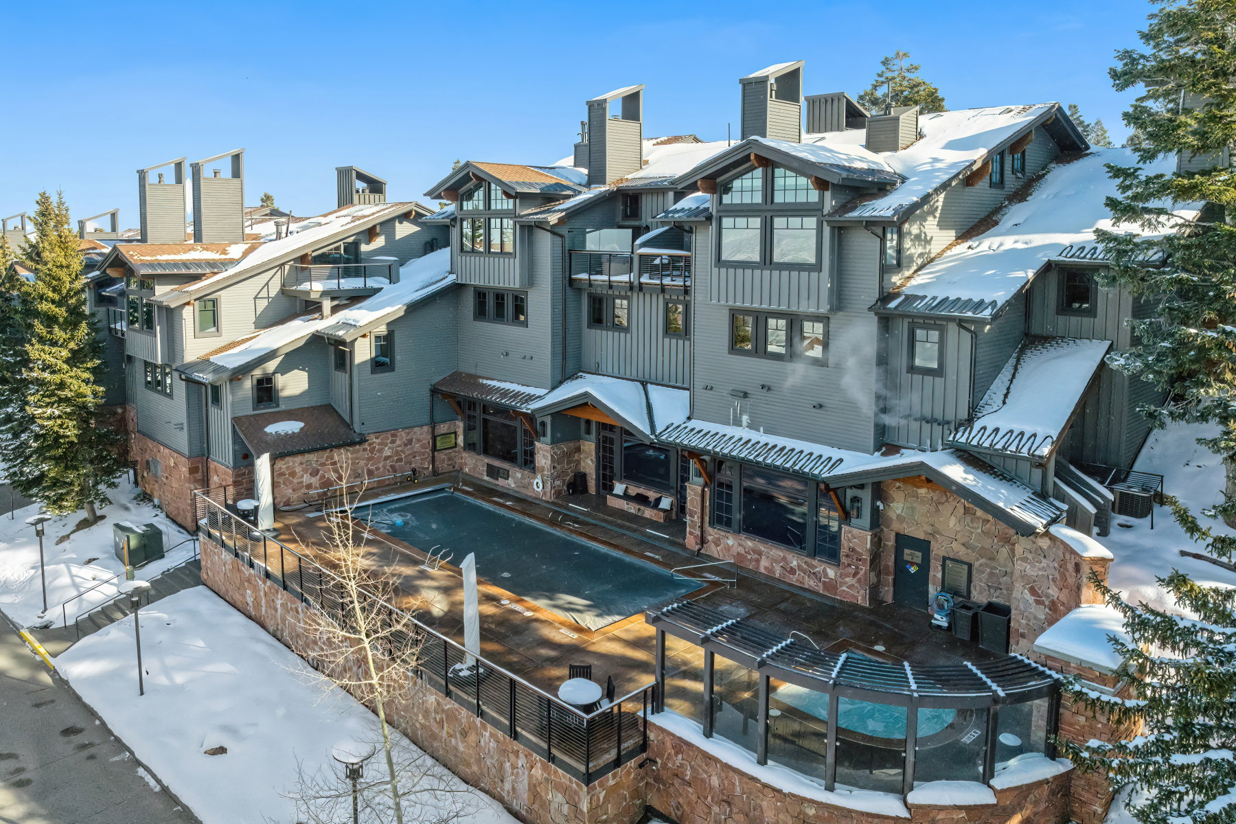 Contemporary multi-story building featuring stone and siding exterior, multiple balconies, an outdoor swimming pool, and an outdoor hot tub with a pergola