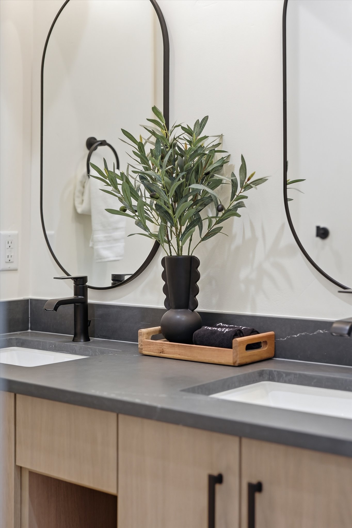 Double vanity featuring a dark countertop with integrated sinks, matte black faucets, wood-finish cabinetry, and contemporary oval mirrors