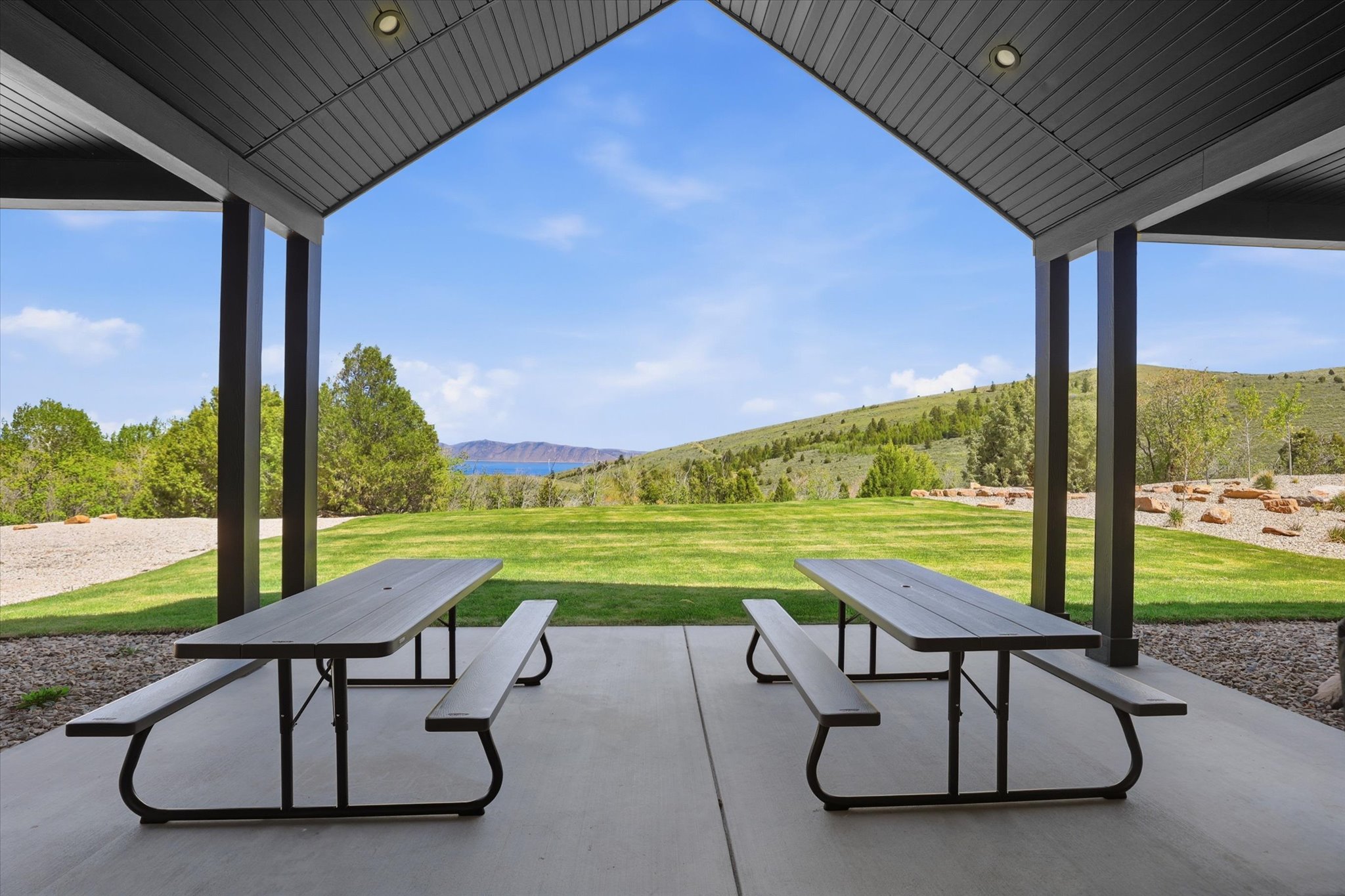 Covered patio area with concrete flooring and dark support beams, offering expansive lawn and distant water body