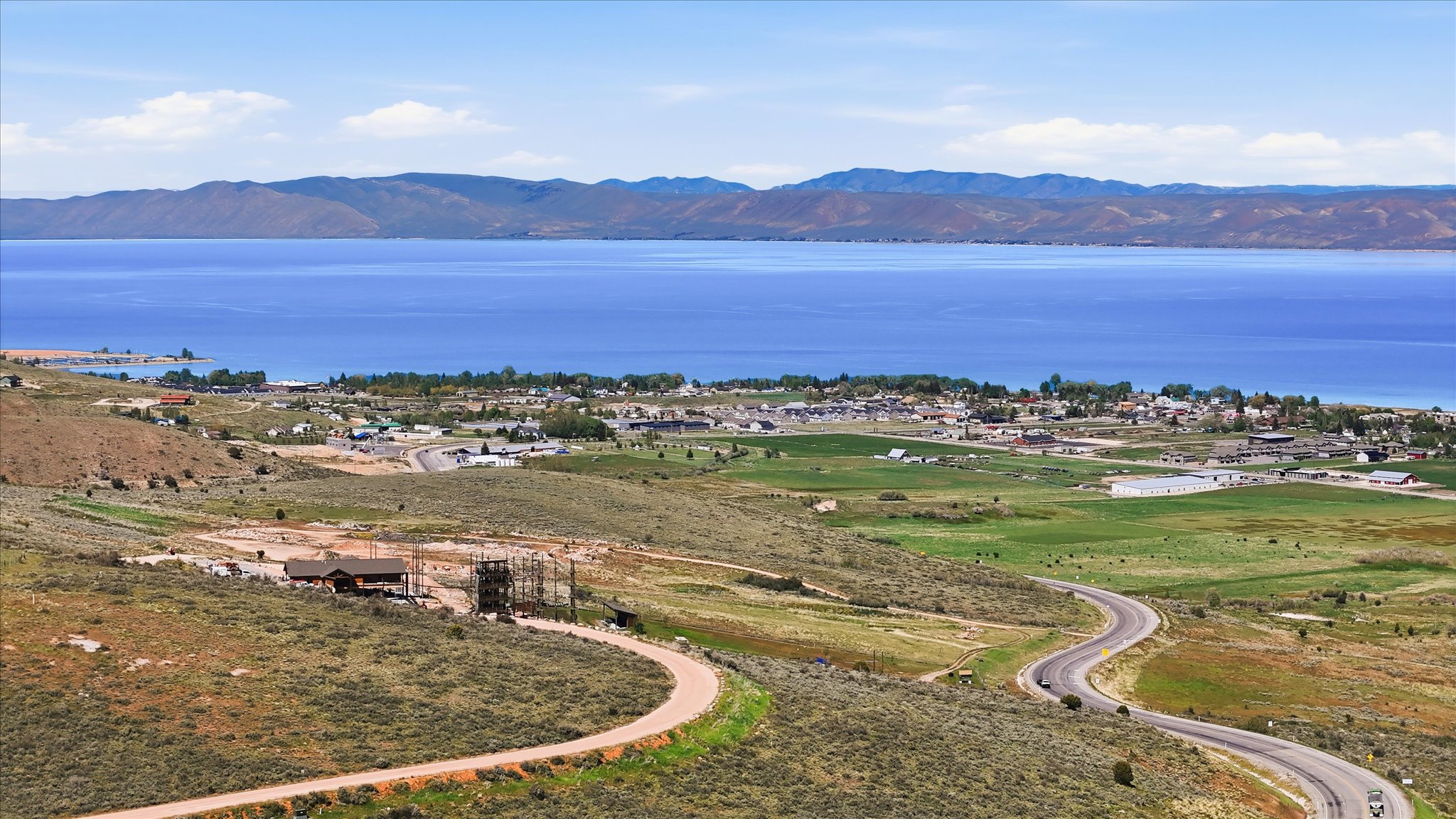 Expansive property featuring a winding paved road, a partially developed building site, and a background of a large body of water