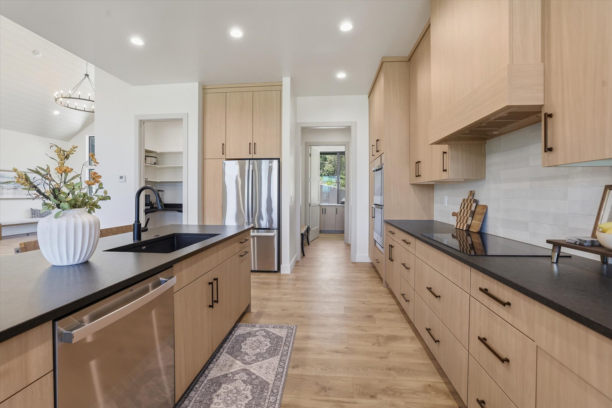 Contemporary kitchen featuring extensive light wood-finish cabinetry, dark countertops, integrated stainless steel appliances, a large island with an undermount sink and matte black faucet, and durable wood-finish flooring