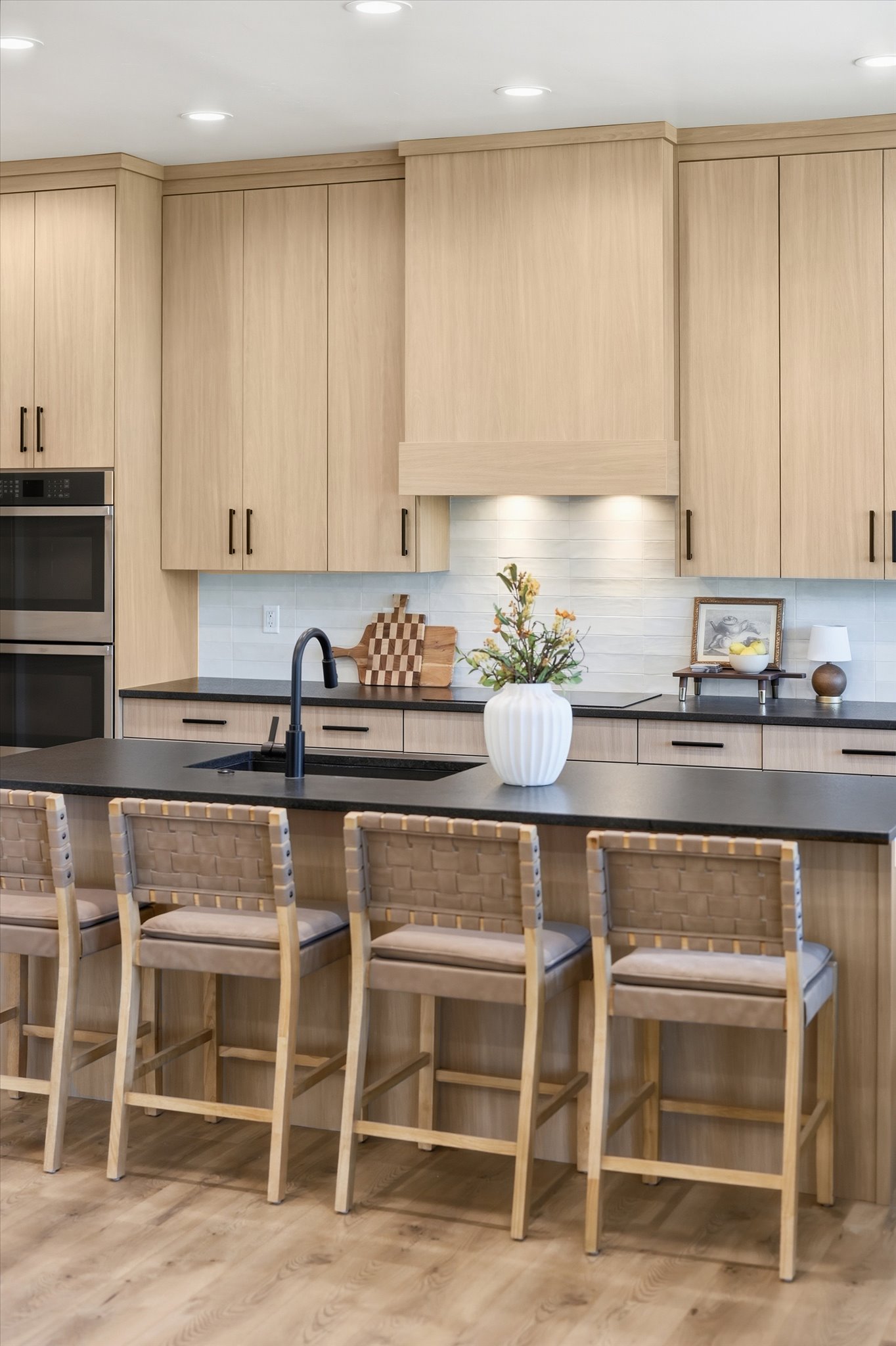 Kitchen featuring light wood-finish cabinetry with black hardware, a black countertop island with integrated sink, recessed ceiling lighting, a white subway tile backsplash, and built-in stainless steel double ovens
