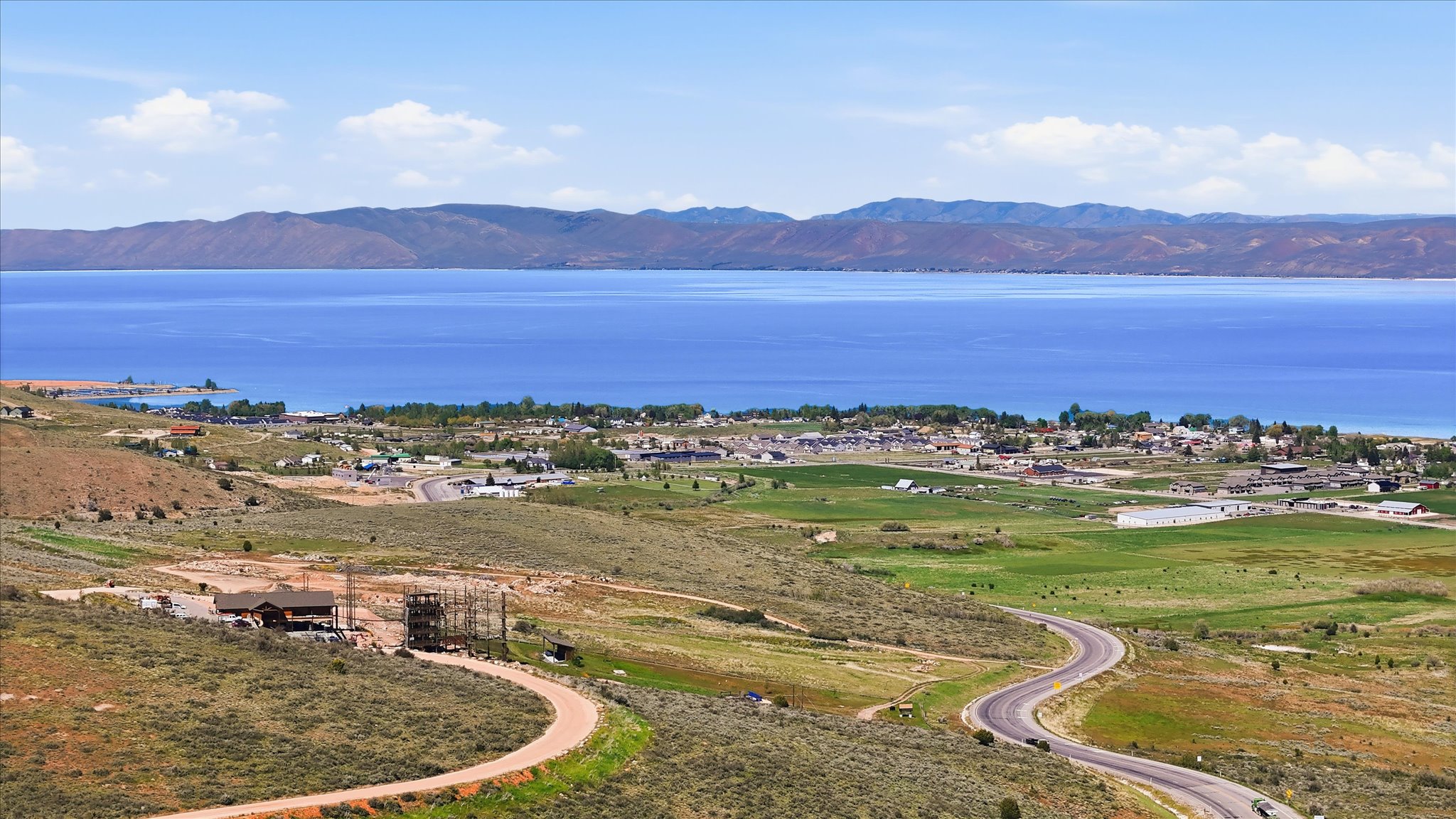 Expansive aerial landscape featuring a large body of water, distant mountain ranges, a small community along the shoreline, and verdant fields