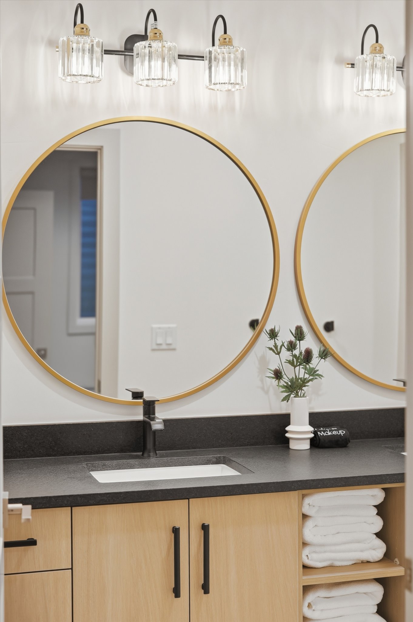 Dual circular mirrors with gold-finish frames, a three-light vanity fixture featuring fluted glass shades, a dark countertop with an integrated rectangular sink, light wood-finish cabinetry, and matte black hardware
