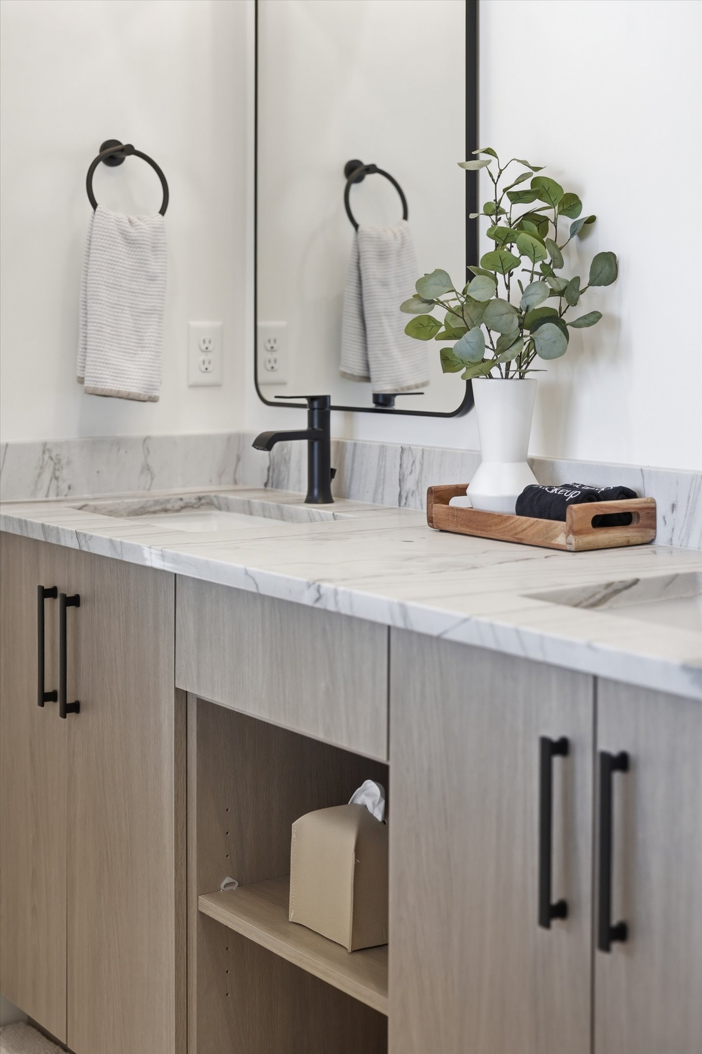 Dual vanity with a stone countertop, undermount sinks, wood-finish cabinetry, matte black hardware, and a framed mirror