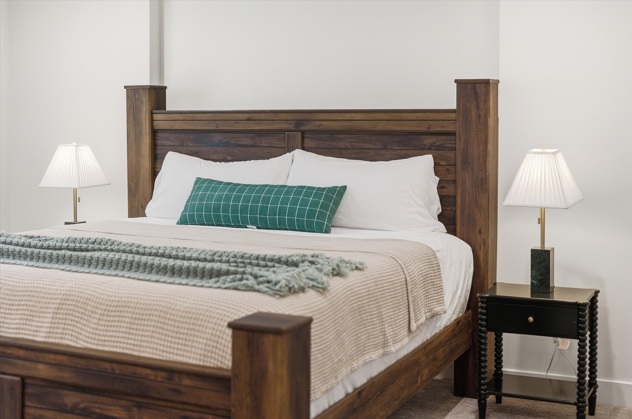 Spacious sleeping area featuring a robust wood-finish bed frame, light-colored walls, and a contrasting dark side table with a contemporary lamp