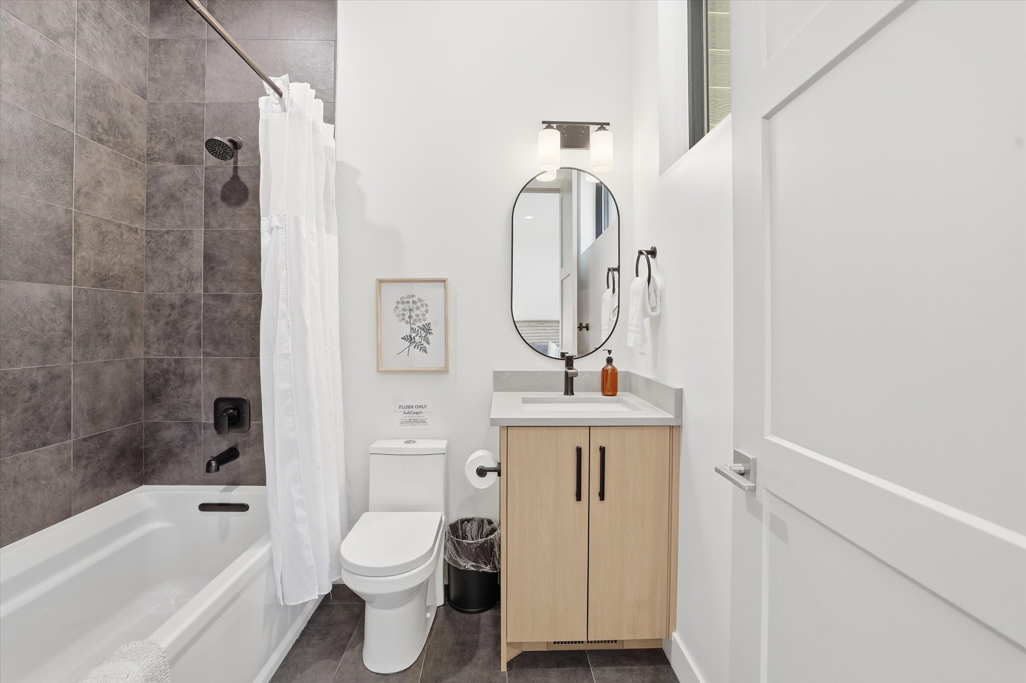 Contemporary full bathroom featuring a tub/shower combination with dark large-format wall tiles, a light wood-finish vanity with a light-toned countertop, a black-framed oval mirror, and a modern light fixture