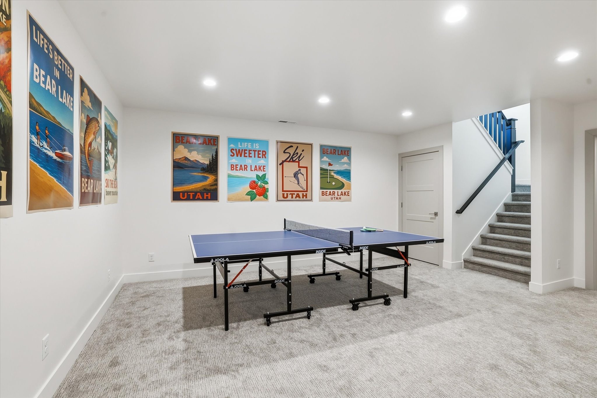 Spacious room featuring light-colored carpet, recessed lighting, a white door, and a staircase with a dark railing