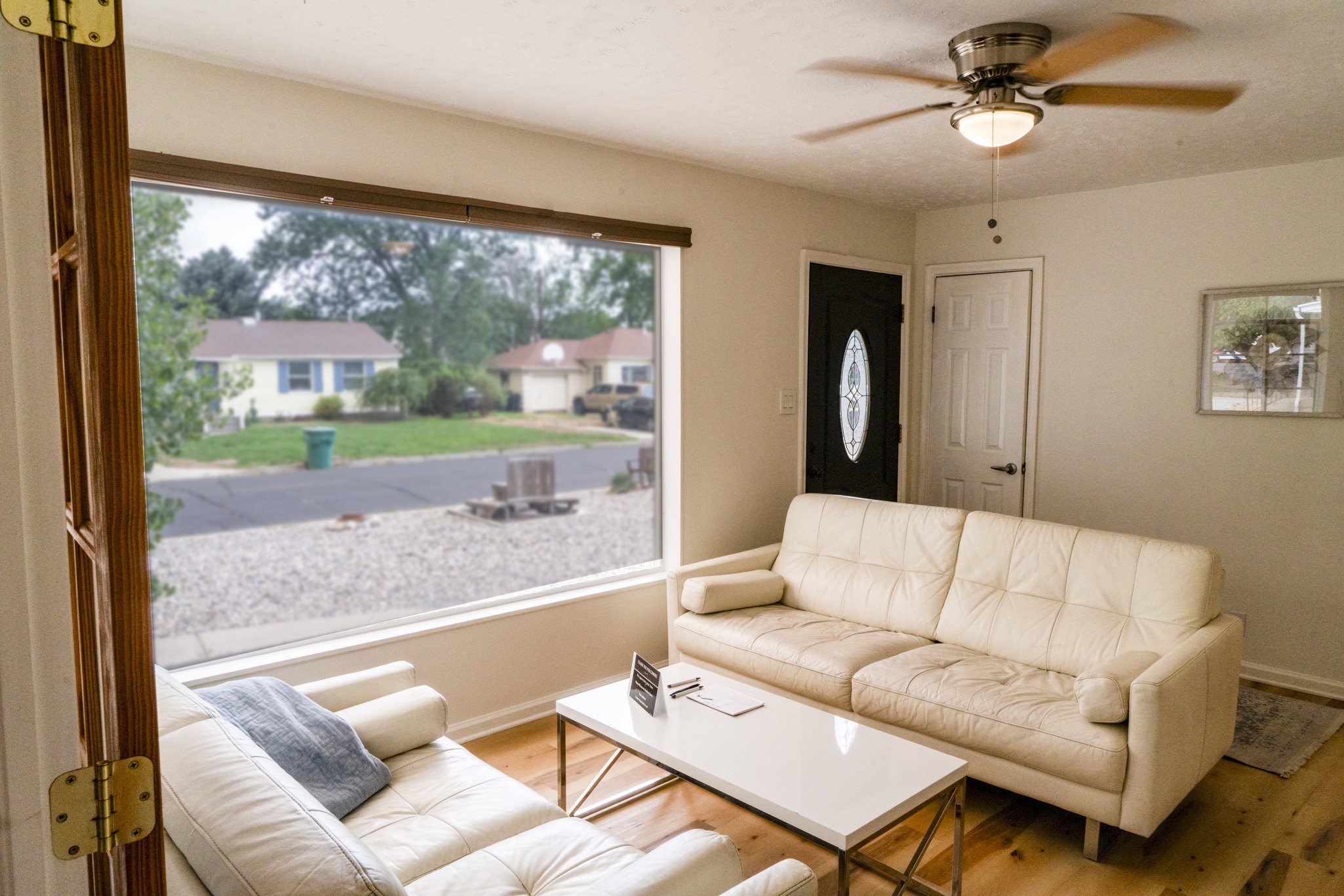Living area featuring a large picture window, wood-finish flooring, a ceiling fan with light fixture, and a front entry door with an oval glass insert