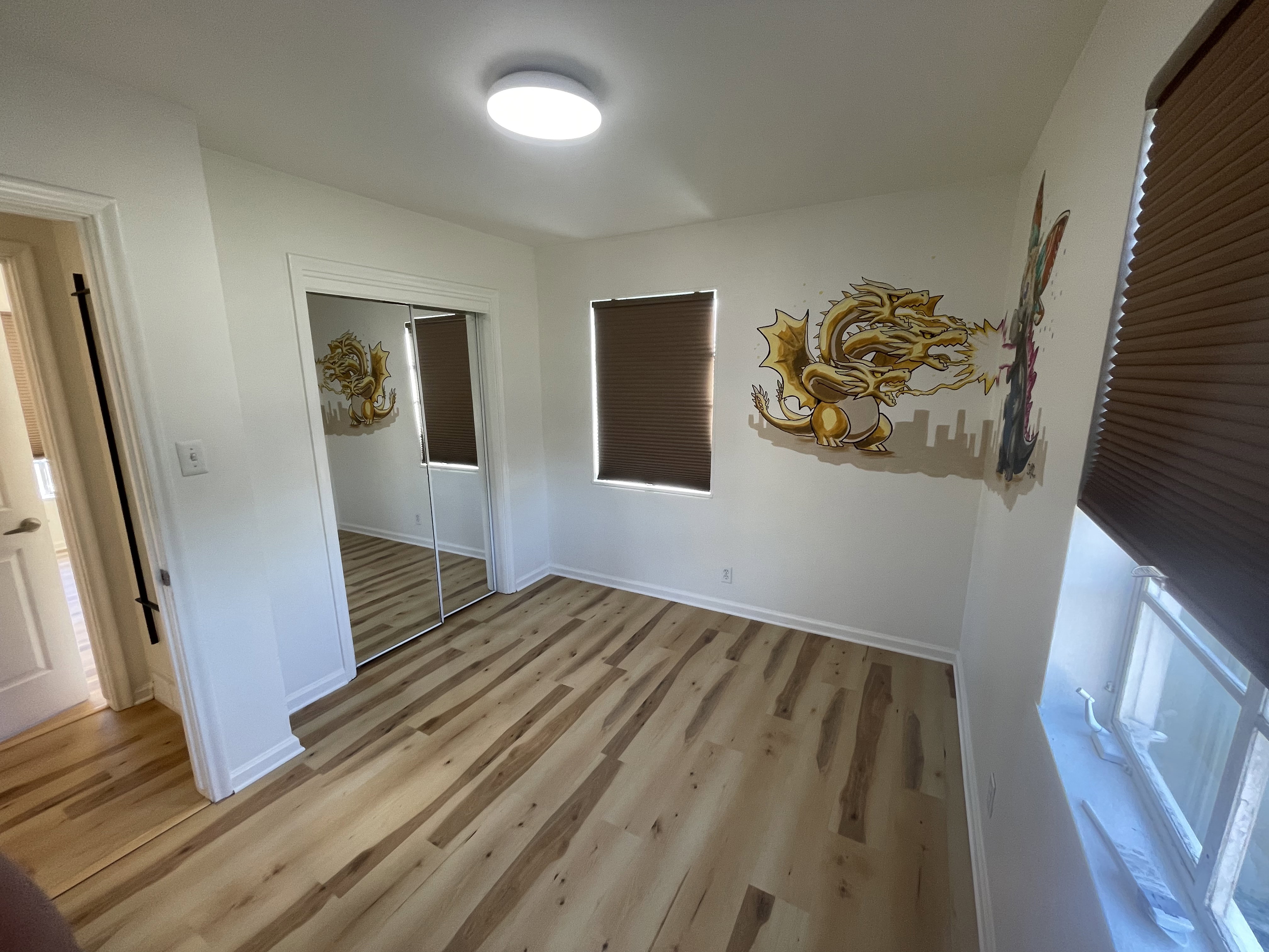 Light-filled room featuring wood-finish flooring, a full-length mirrored closet, two windows with brown blinds, and a flush-mount ceiling light fixture