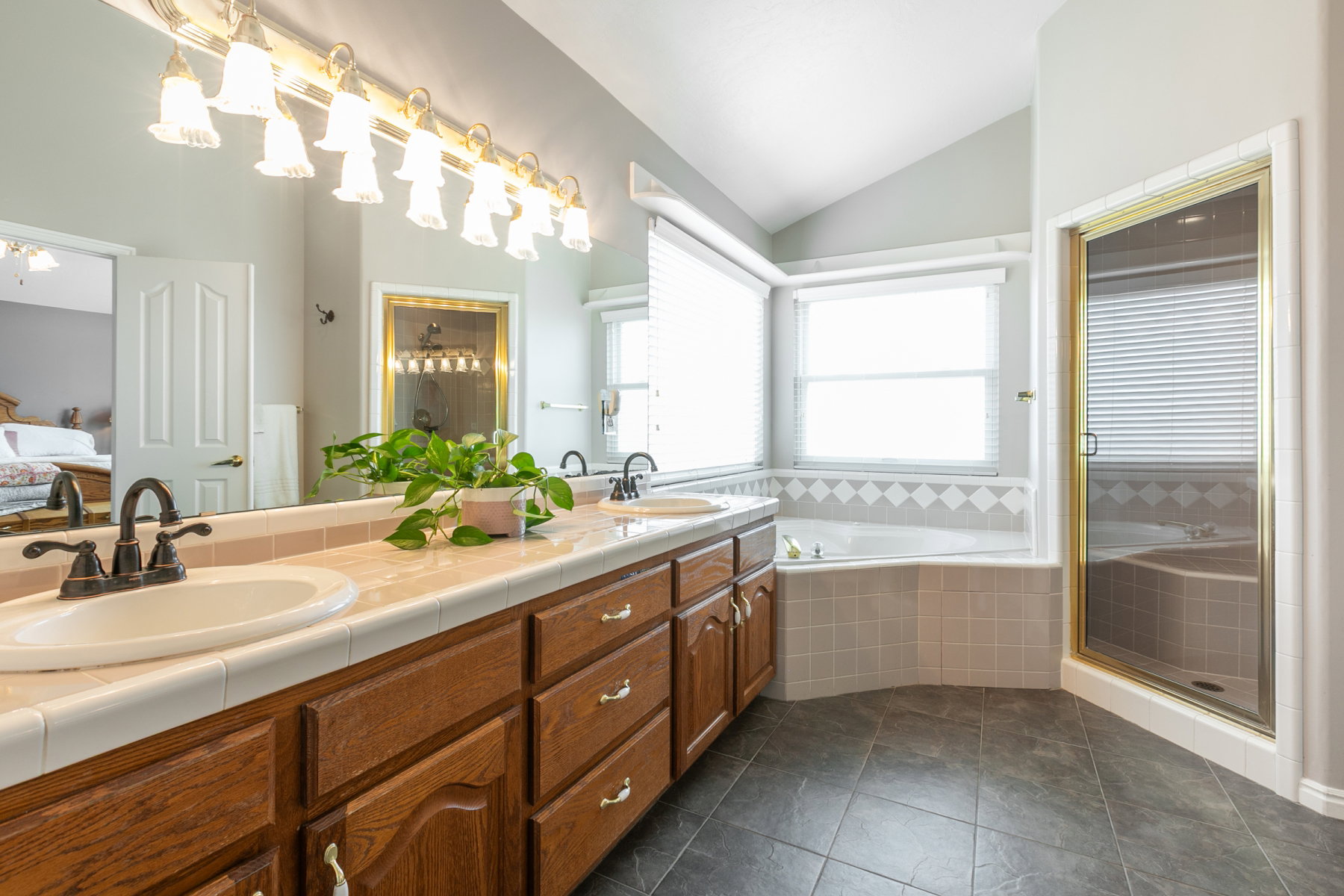 Spacious bathroom featuring a dual vanity with a tile countertop, a large soaking tub with tile surround, a separate shower enclosure with a gold-toned frame, and dark tile flooring