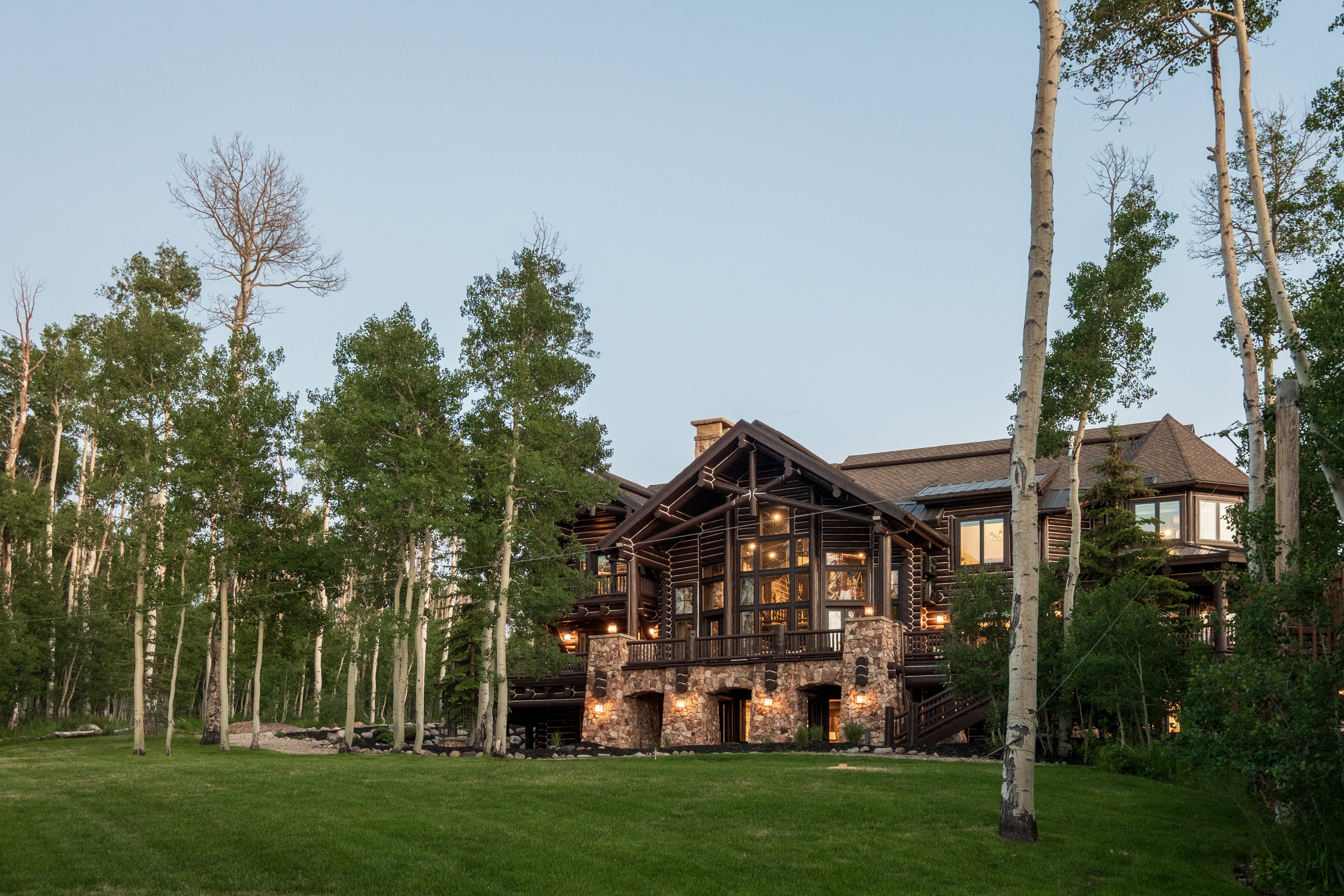 Rustic-style residence featuring a stone foundation, wood-beam construction, expansive multi-pane windows, multiple exterior lighting fixtures, and a spacious green lawn