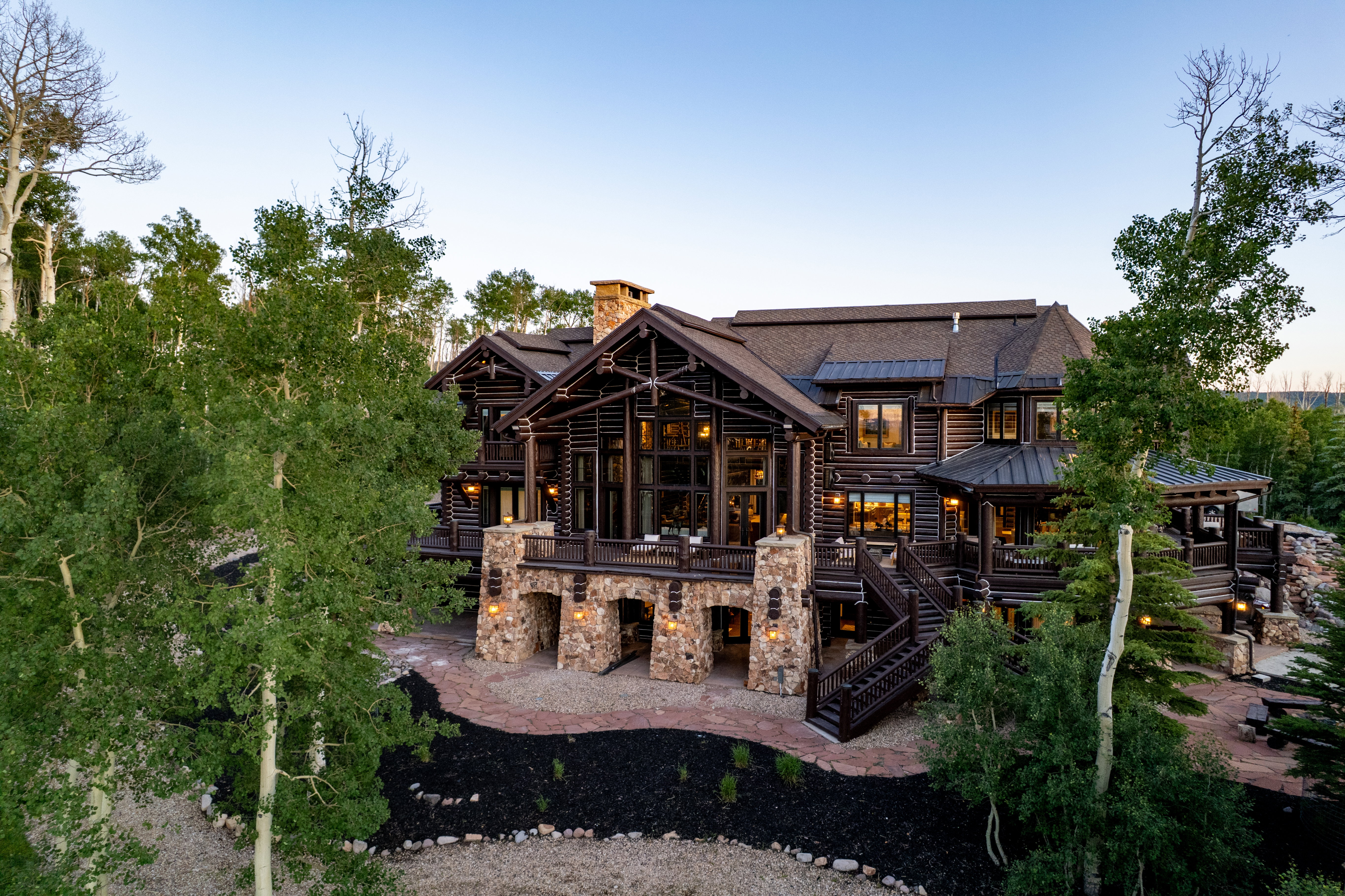 Multi-level log cabin exterior featuring a stone foundation, prominent stone chimney, multiple outdoor decks, and extensive window arrays