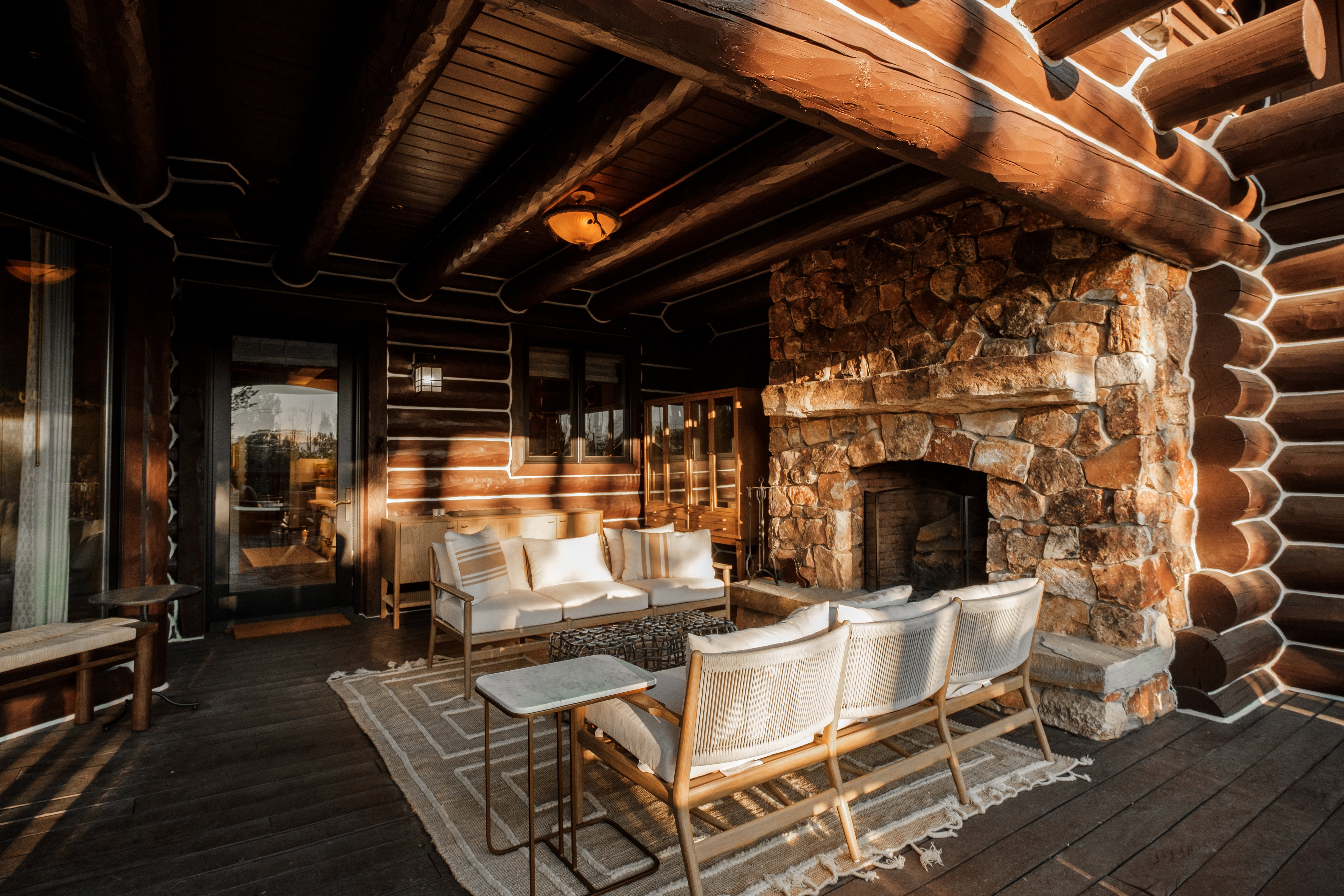 Covered outdoor living space featuring a stone fireplace, exposed log beams, wood-finish flooring, and a glass-paneled door