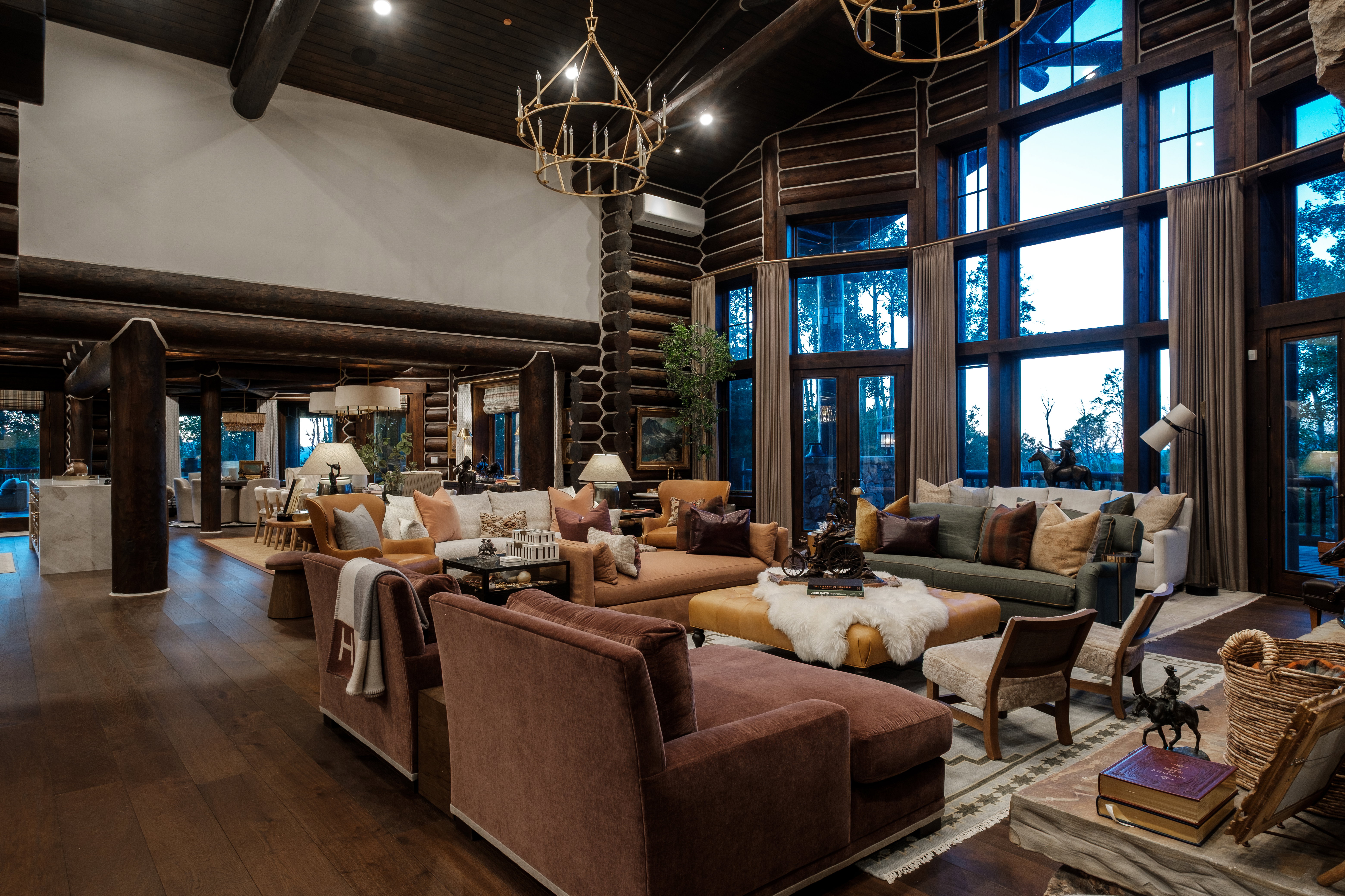 Expansive great room featuring a grand vaulted ceiling, floor-to-ceiling windows, exposed log beams, wood-finish flooring, and two brass chandeliers