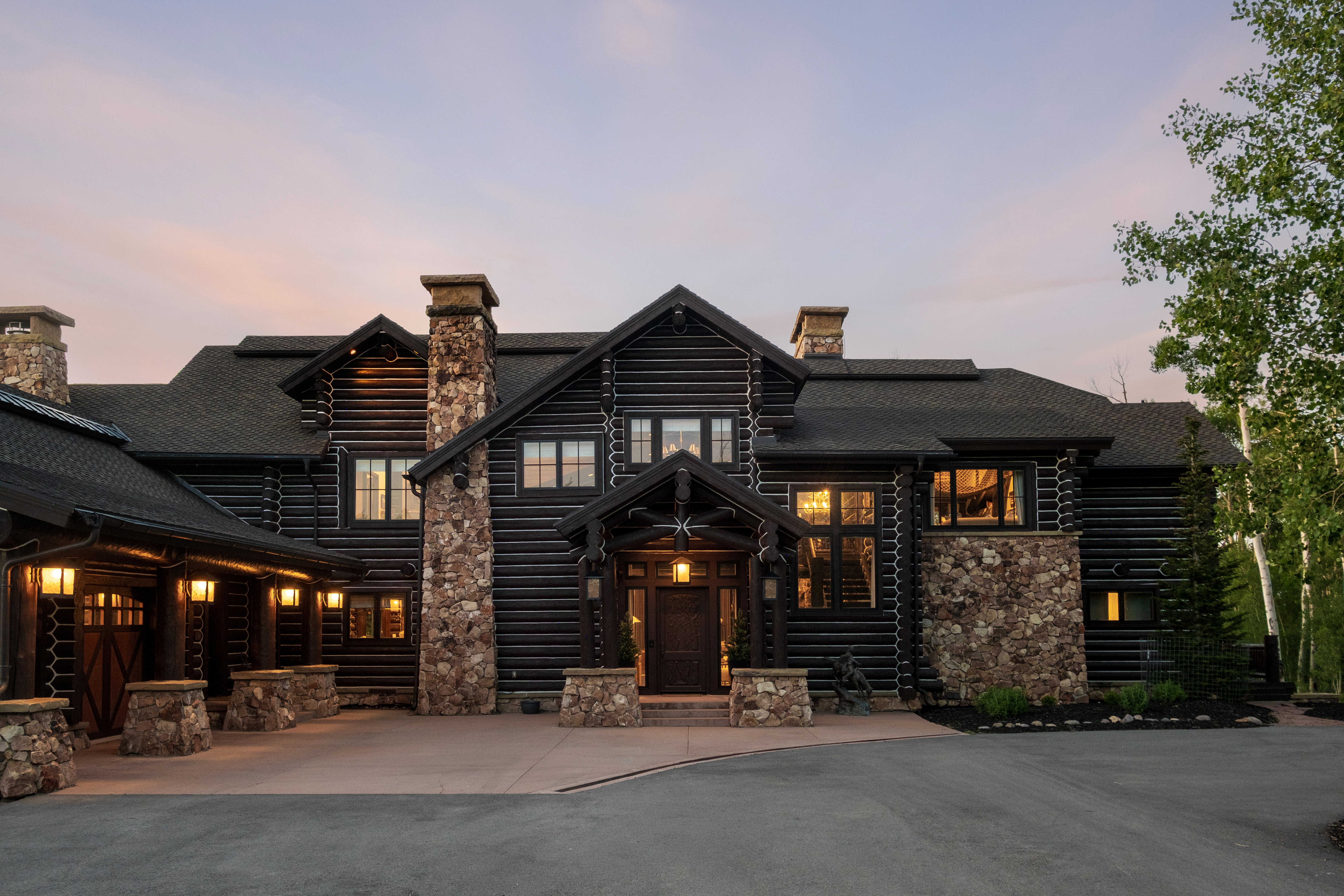Striking log home with prominent stone chimneys, a covered entryway, a connected garage with carriage-style doors, and asphalt shingle roofing