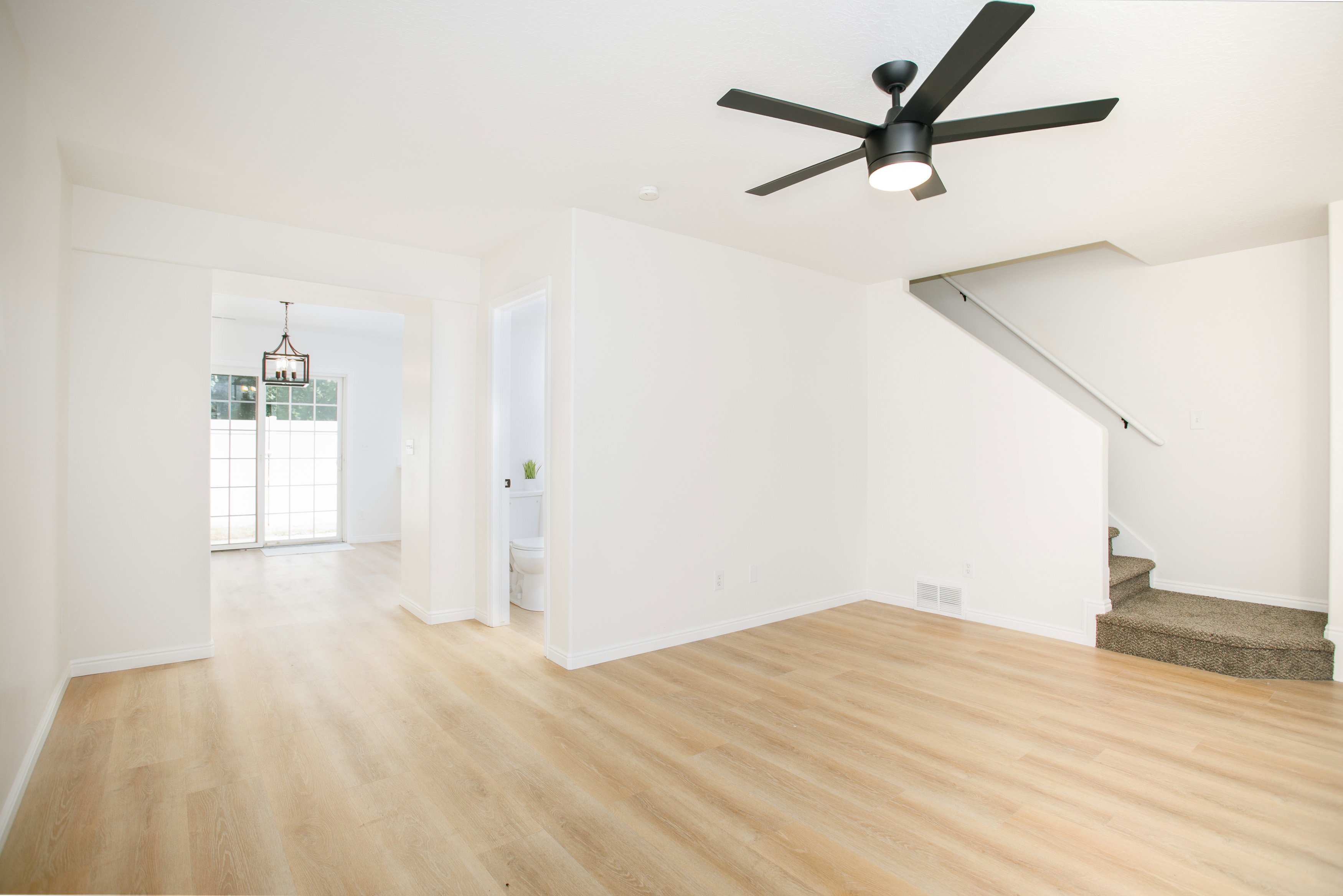 Spacious living area featuring light wood-finish flooring, a modern black ceiling fan, and white walls