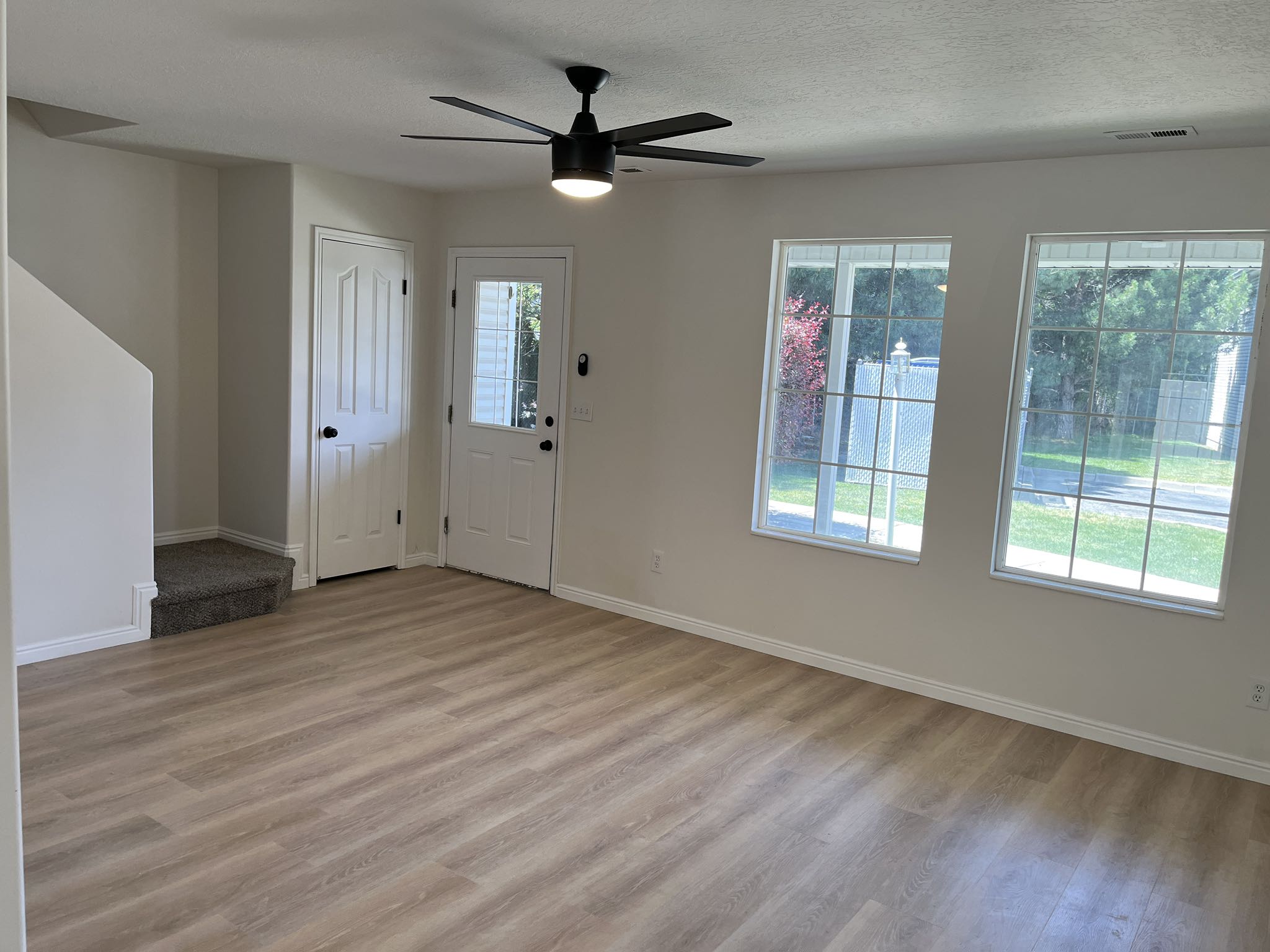 Spacious room featuring wood-finish flooring, two large windows, a ceiling fan with integrated lighting, a white paneled door, and a white exterior door with a glass insert