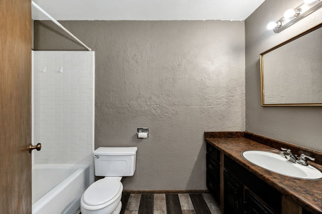 Bathroom featuring a white tub and shower surround with tile, a vanity with a white oval sink, a brown patterned countertop, dark wood-finish cabinetry, and wood-finish flooring