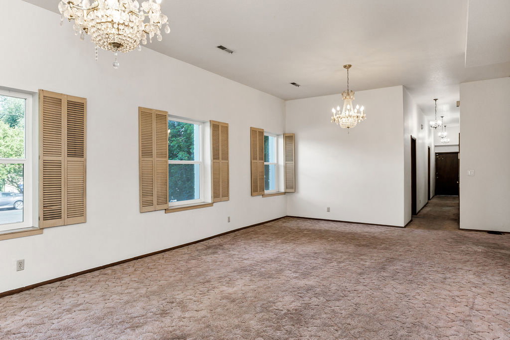 Spacious interior room featuring two crystal chandeliers, three windows with natural wood louvered shutters, and textured wall-to-wall carpeting