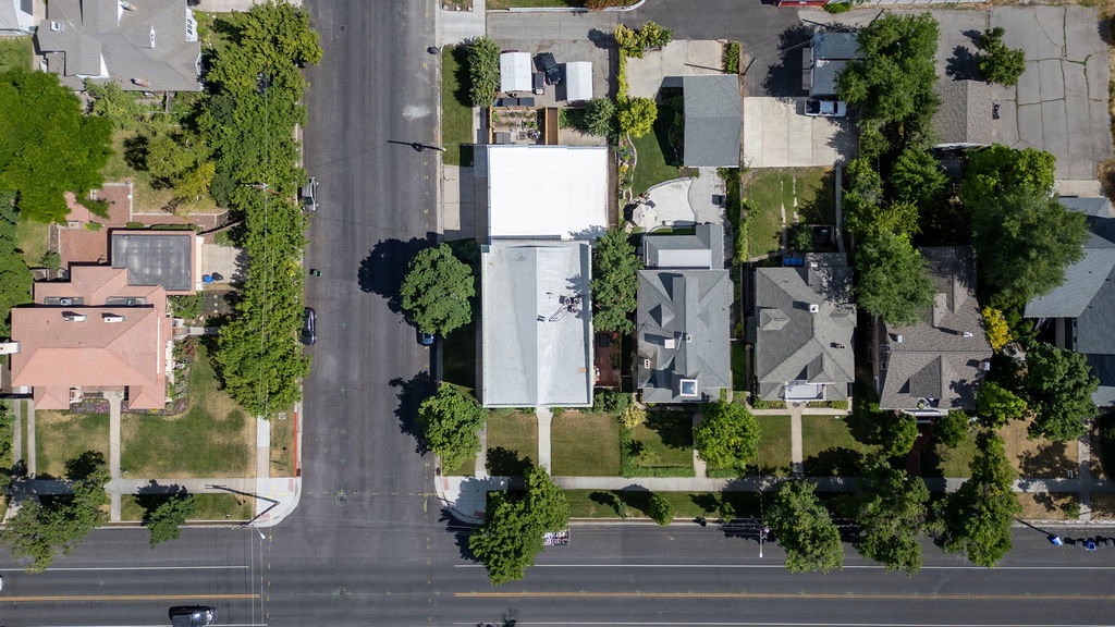 Aerial perspective showcasing a residential property with a light-colored roof, surrounded by mature trees and paved surfaces