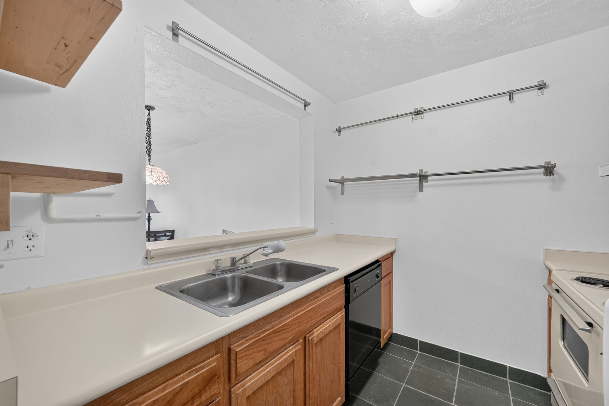 Kitchen featuring a double basin stainless steel sink, light wood-finish cabinetry, a built-in black dishwasher, dark tile flooring, and a pass-through window