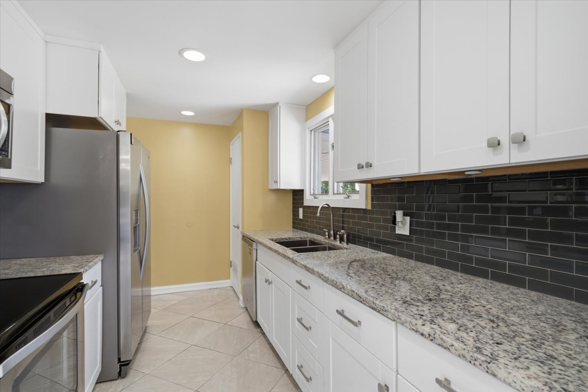 Galley kitchen featuring white cabinetry, light-toned granite countertops, a dark subway tile backsplash, stainless steel appliances, and recessed lighting