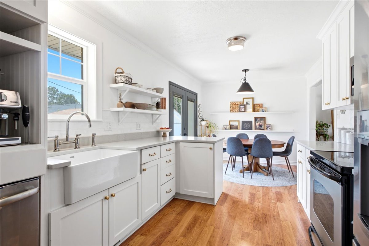 White kitchen featuring a farmhouse sink, stainless steel appliances, wood-finish flooring, white cabinetry with brass hardware, and an adjacent dining area