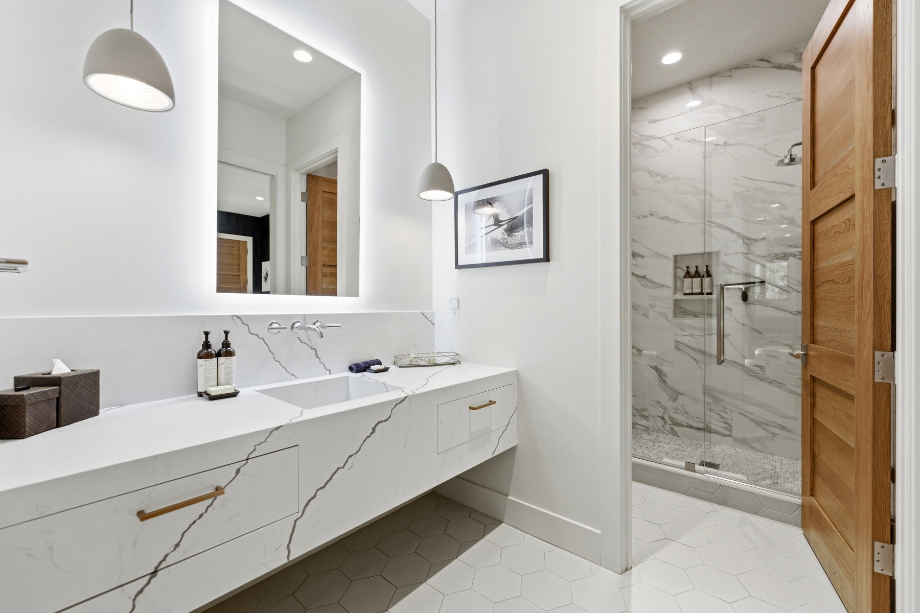 Bathroom featuring a floating double vanity with an integrated rectangular sink and wall-mounted faucet, hexagon floor tiles, illuminated mirror, and a glass-enclosed shower with full-height marble-look tile surround and a tiled niche